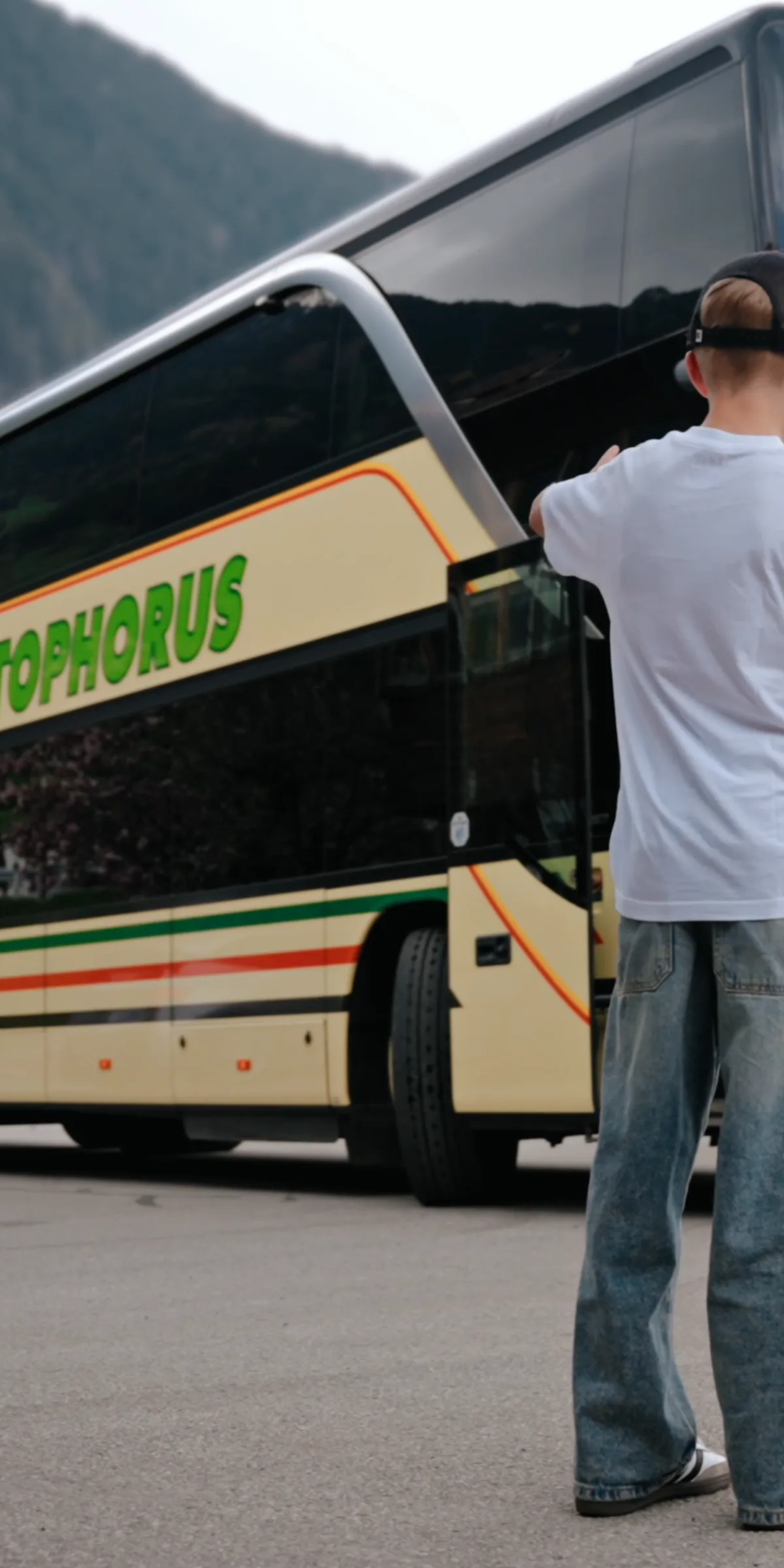 Ein Junge mit Basecap steht auf einem Parkplatz vor einem Reisebus mit dem Schriftzug Christophorus, dahinter ein zweiter Bus und die bewaldeten Hänge des Zillertals.