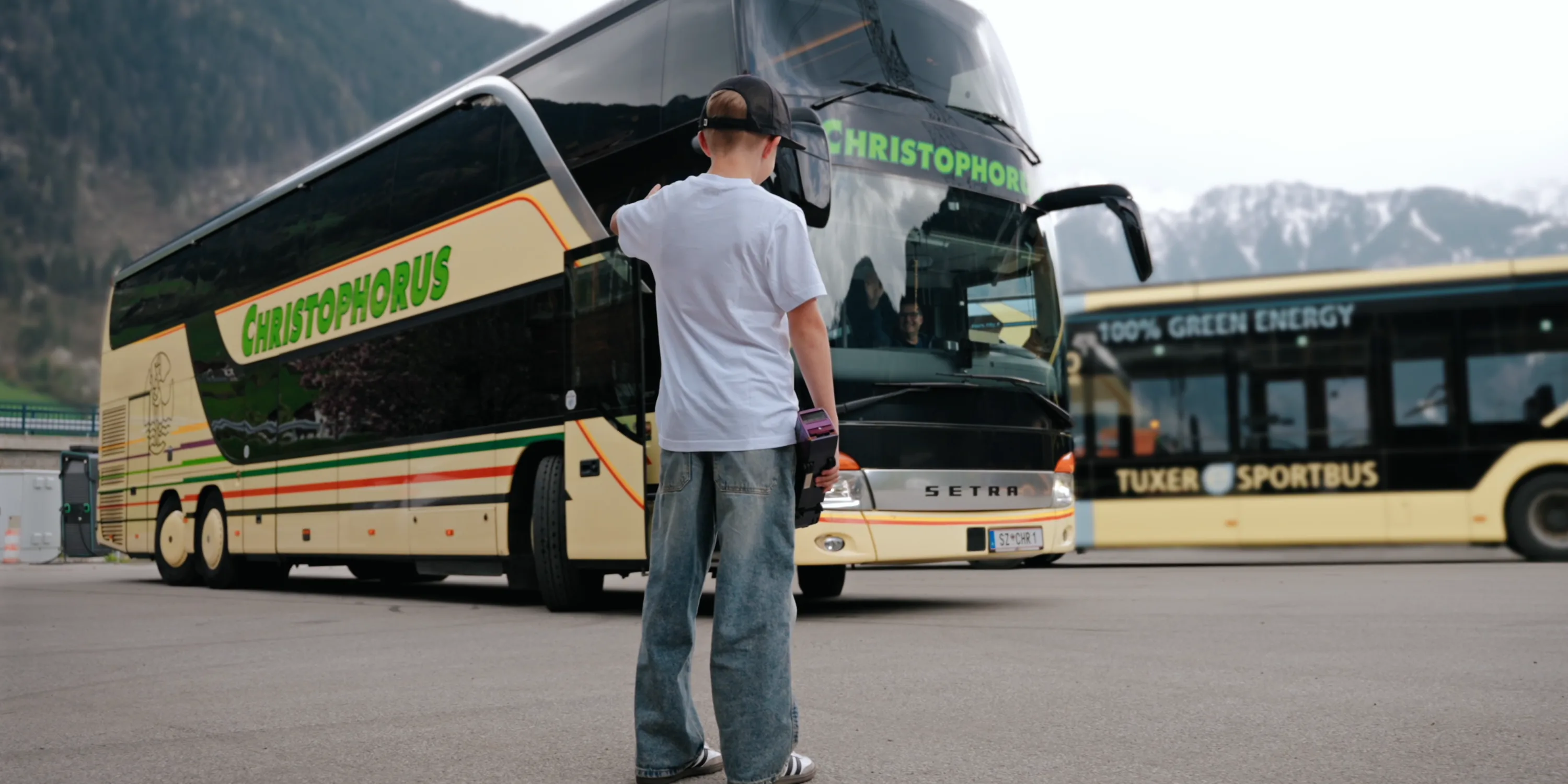 Christophorus Reisen: Ein Junge mit Basecap steht auf einem Parkplatz vor einem Reisebus mit dem Schriftzug Christophorus, dahinter ein zweiter Bus und die bewaldeten Hänge des Zillertals.