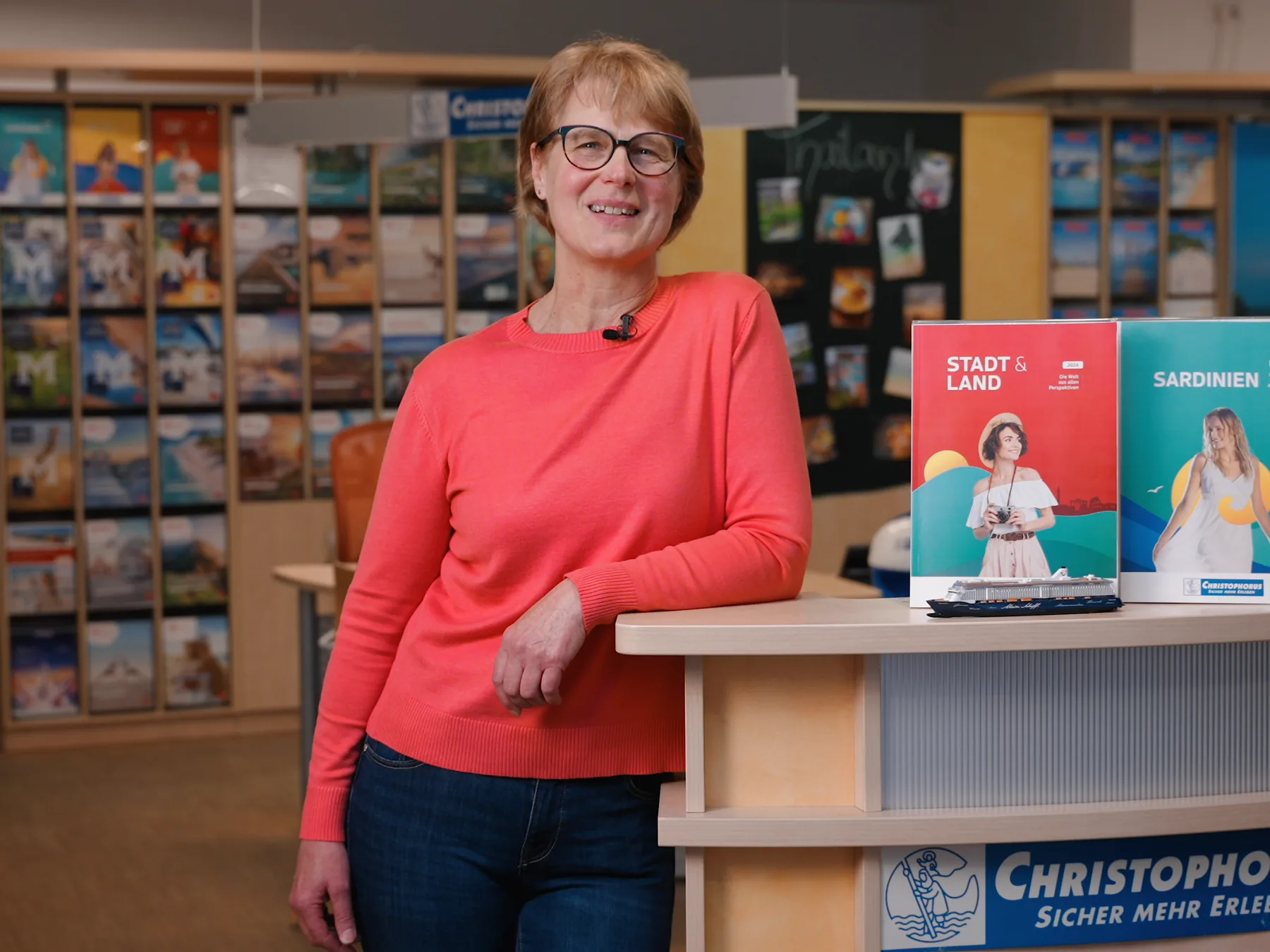 Eine Beraterin im roten Pullover lehnt an der Theke eines Christophorus-Reisebüros, hinter ihr eine Wand voller Reisekataloge, vor ihr zwei Prospekte.