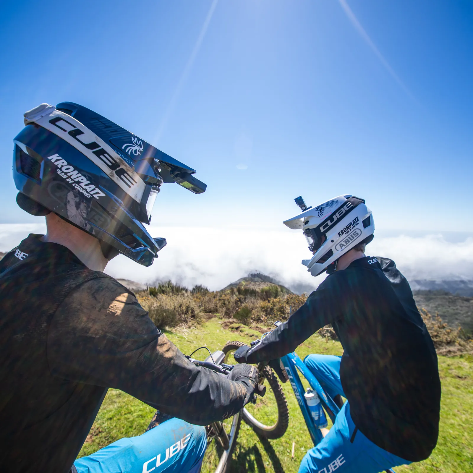Zwei Fahrer in Teamkleidung stehen im Gegenlicht über einem Wolkenmeer, die Helme auf, die Sonne knapp über dem Bildrand.
