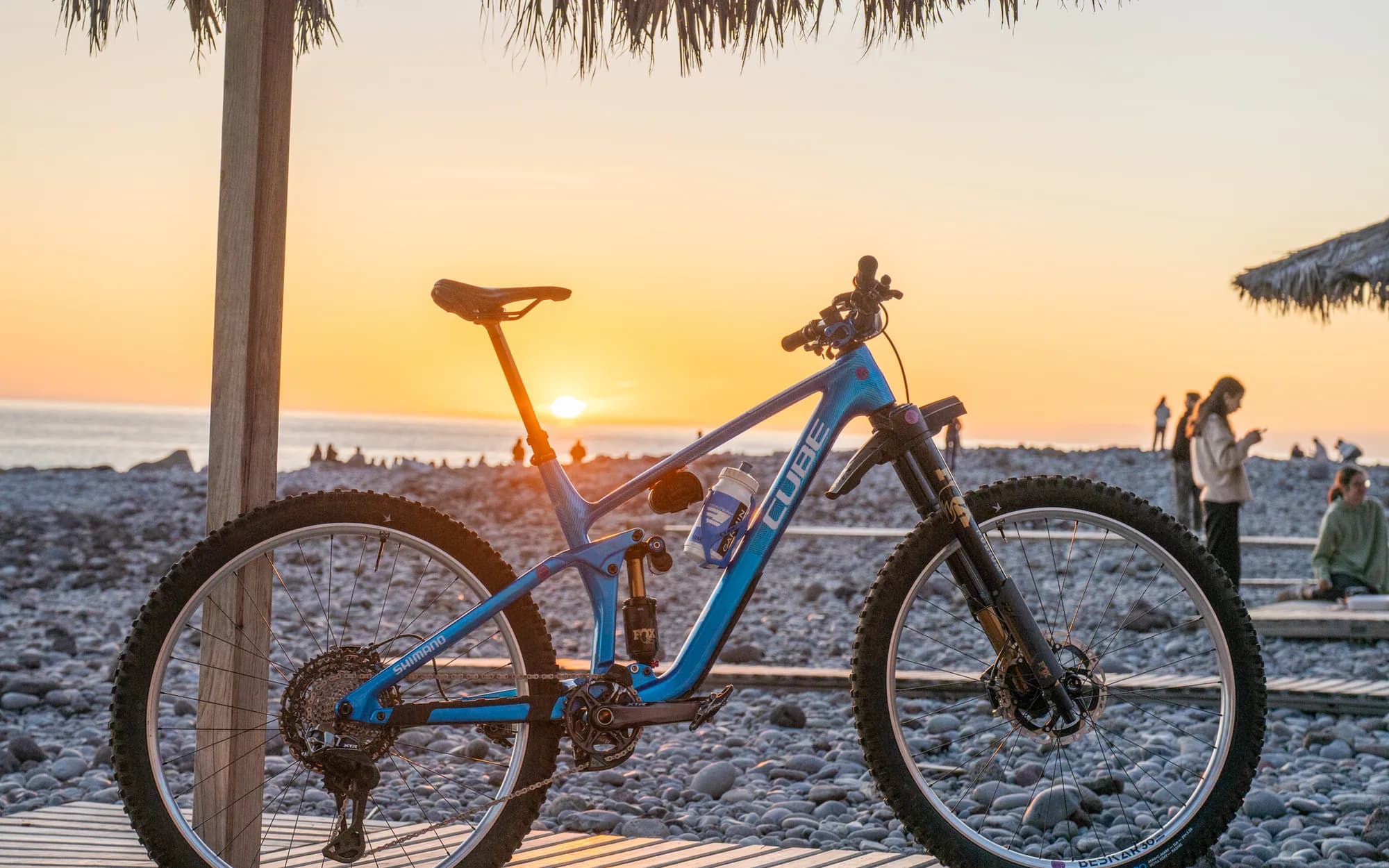 Ein blaues CUBE Mountainbike steht auf einem Holzsteg unter einem Sonnenschirm aus Palmwedeln, dahinter geht die Sonne über dem Meer unter.