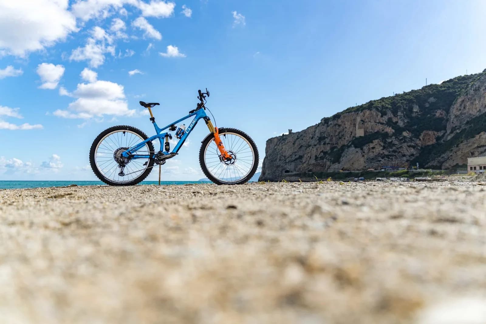 Ein blaues Mountainbike steht allein am Kiesstrand, aufgenommen dicht über dem Boden, dahinter Meer, Felswand und Wolkenhimmel.