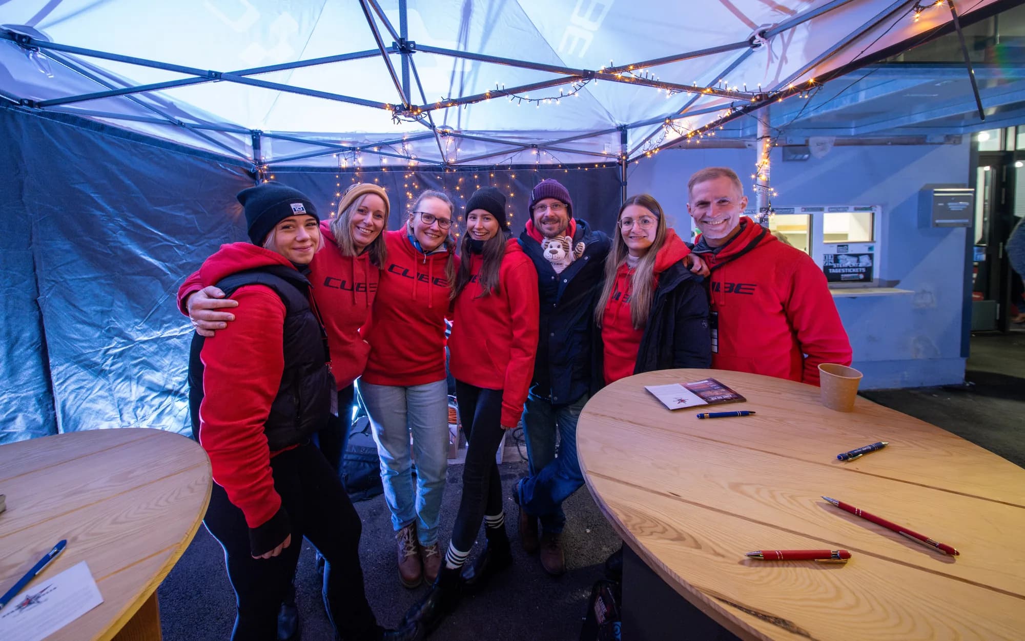 Das Team des CUBE Store Chiemsee in roten Pullovern steht im beleuchteten Zelt vor dem Stadion zusammen, davor runde Stehtische mit Karten und Stiften.