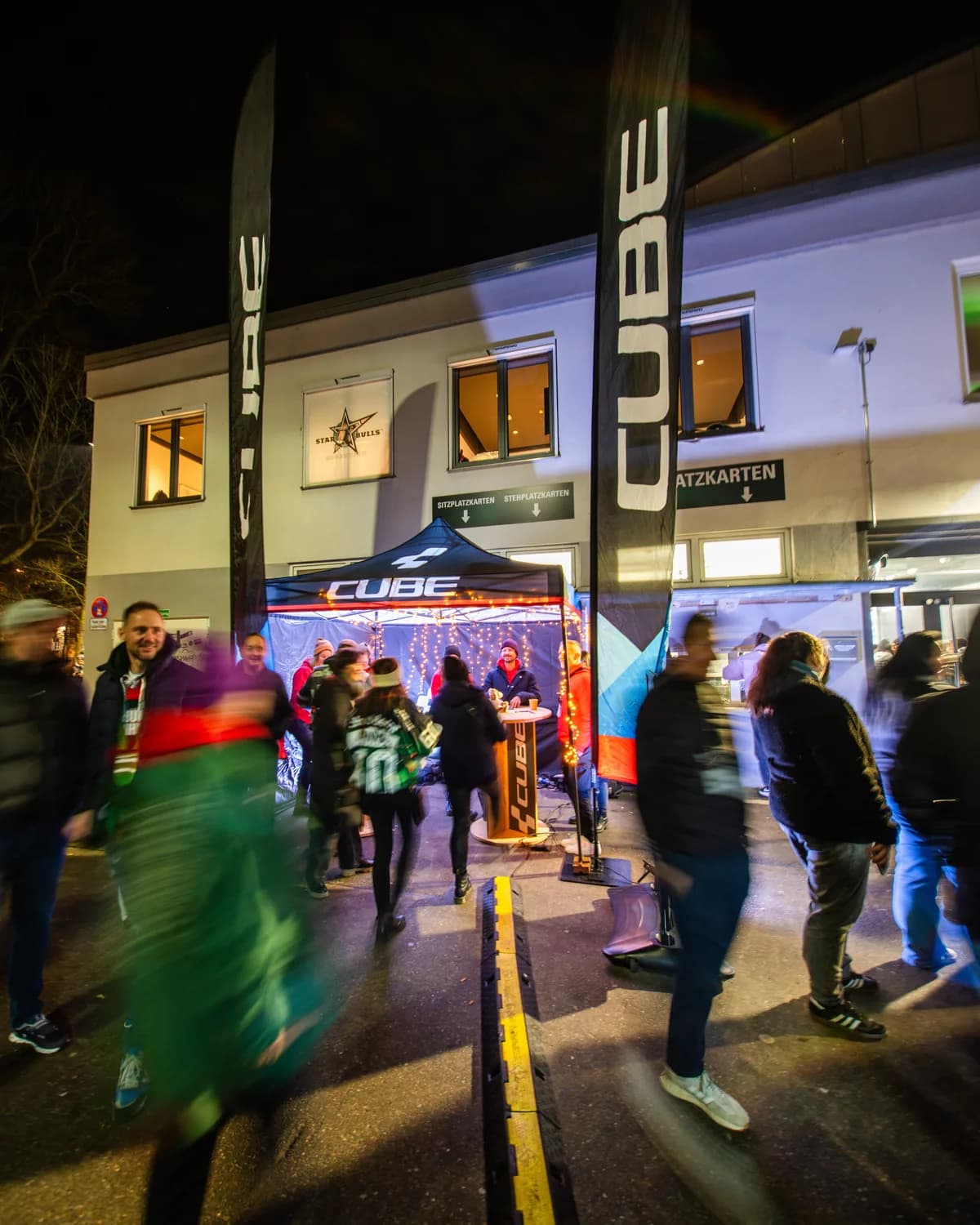 Das Zelt des CUBE Store Chiemsee am Abend vor dem Eingang des Stadions, davor Fans auf dem Weg zum Spiel, daneben eine hohe Fahne mit dem CUBE-Schriftzug.