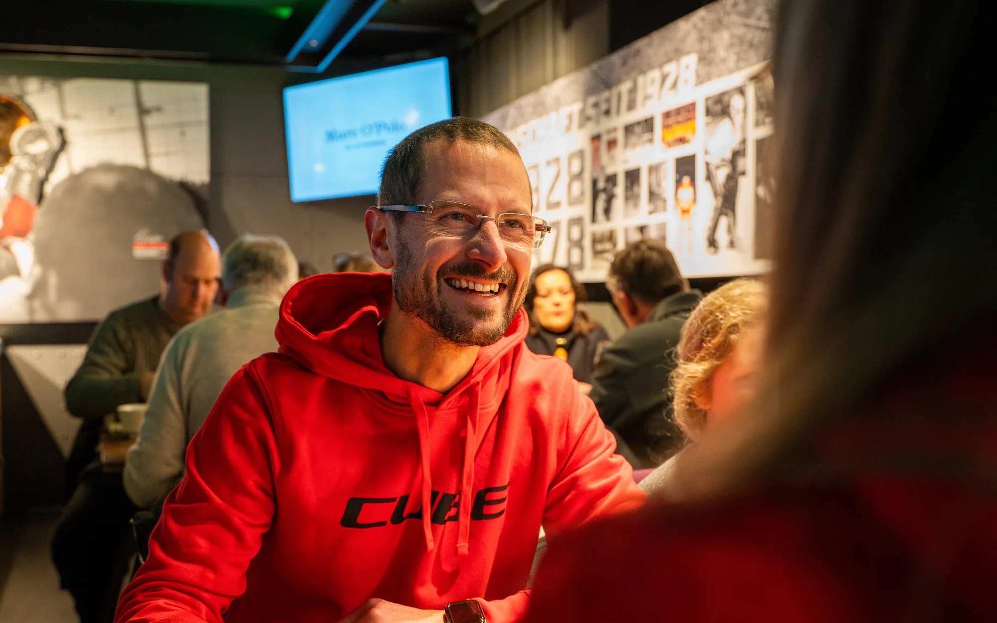 Ein Mann im roten CUBE-Kapuzenpullover sitzt lachend an einem Holztisch im Stadionrestaurant, hinter ihm Gäste und ein Bildschirm an der Wand.