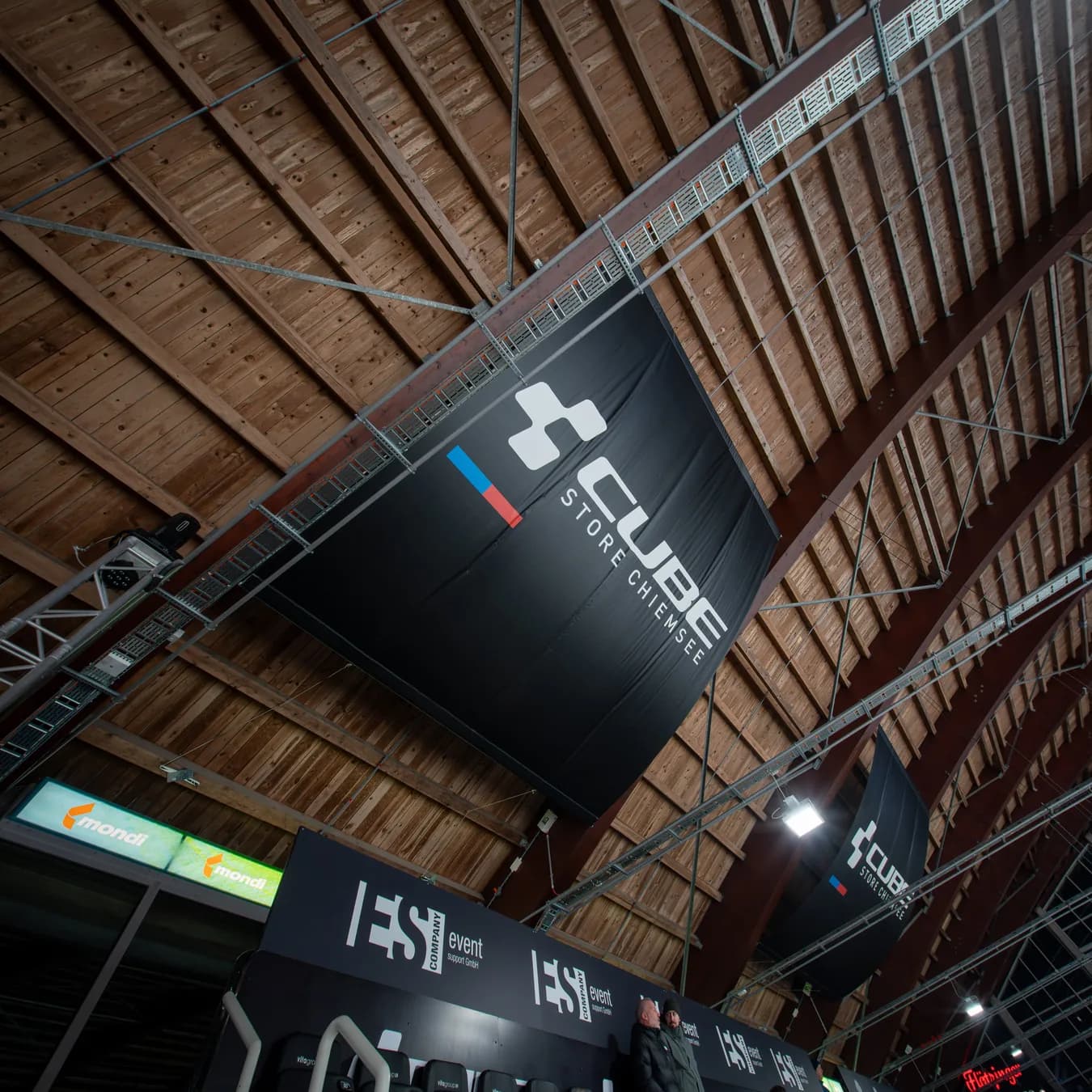 Ein großes schwarzes Banner mit dem Logo des CUBE Store Chiemsee hängt unter dem hölzernen Hallendach des ROFA-Stadions.
