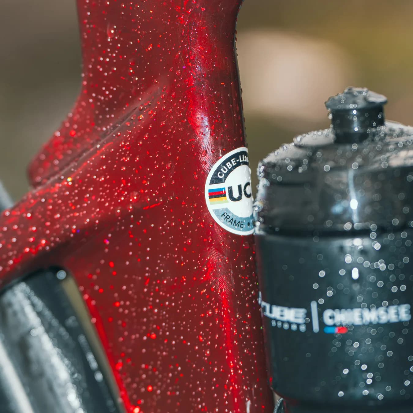 Der glitzernde rote Rahmen mit Wassertropfen, der UCI-Aufkleber und eine schwarze Trinkflasche mit dem Aufdruck CUBE Store Chiemsee.