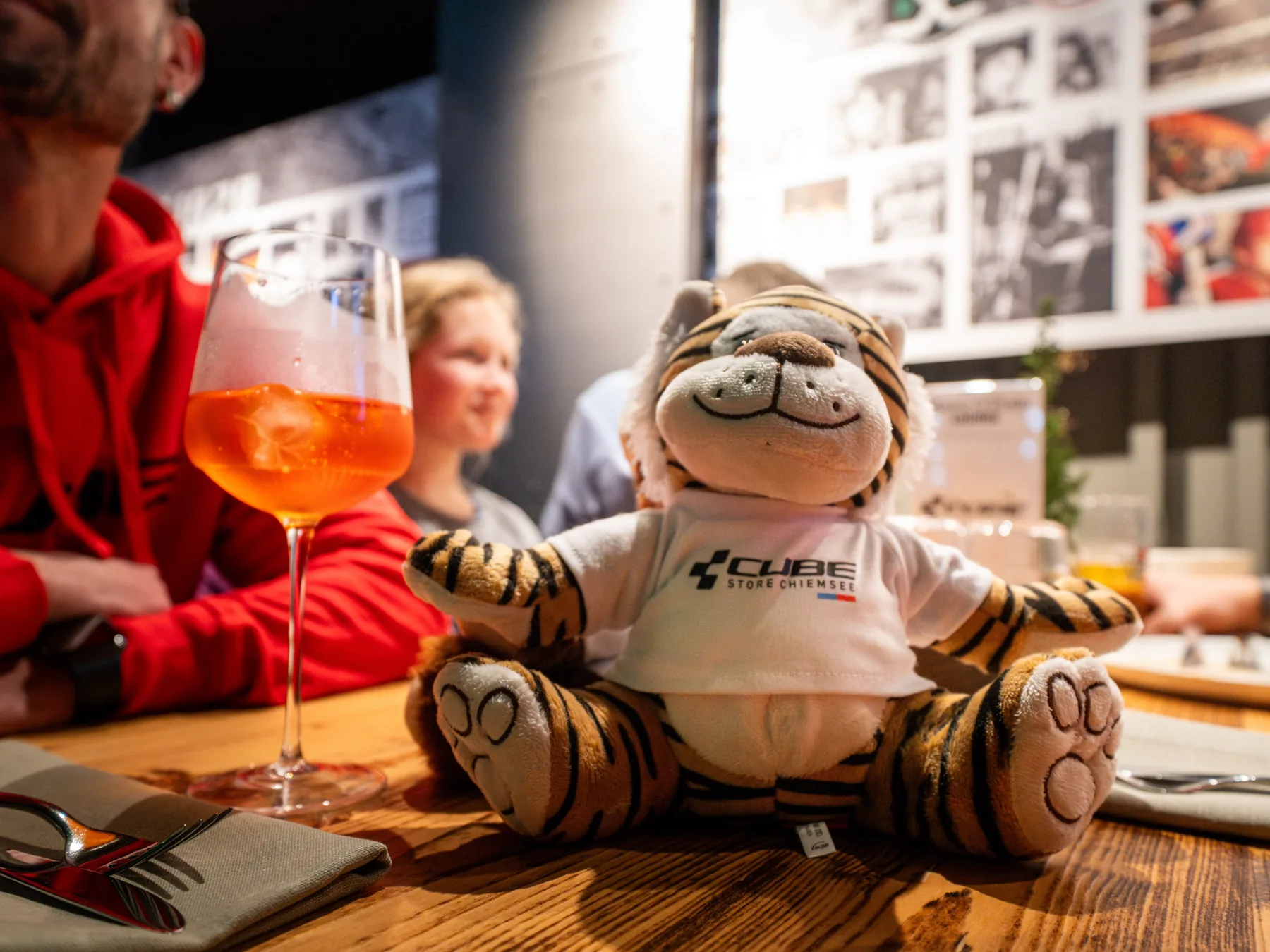 Ein Plüschtiger im weißen Shirt des CUBE Store Chiemsee sitzt auf einem Holztisch im Stadionrestaurant, daneben ein Glas Spritz und Gäste in roten Pullovern.