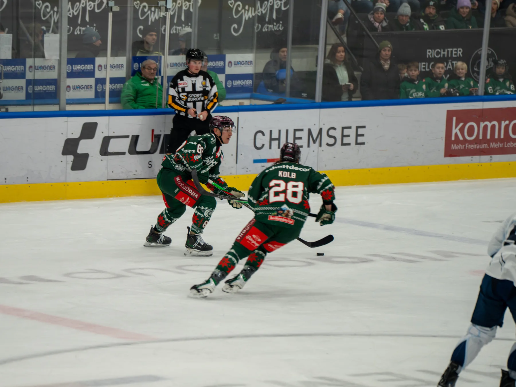 Zwei Spieler der Starbulls Rosenheim in grünen Weihnachtstrikots laufen mit dem Puck vor der Bande mit dem Schriftzug CUBE Chiemsee, dahinter ein Schiedsrichter und Zuschauer.