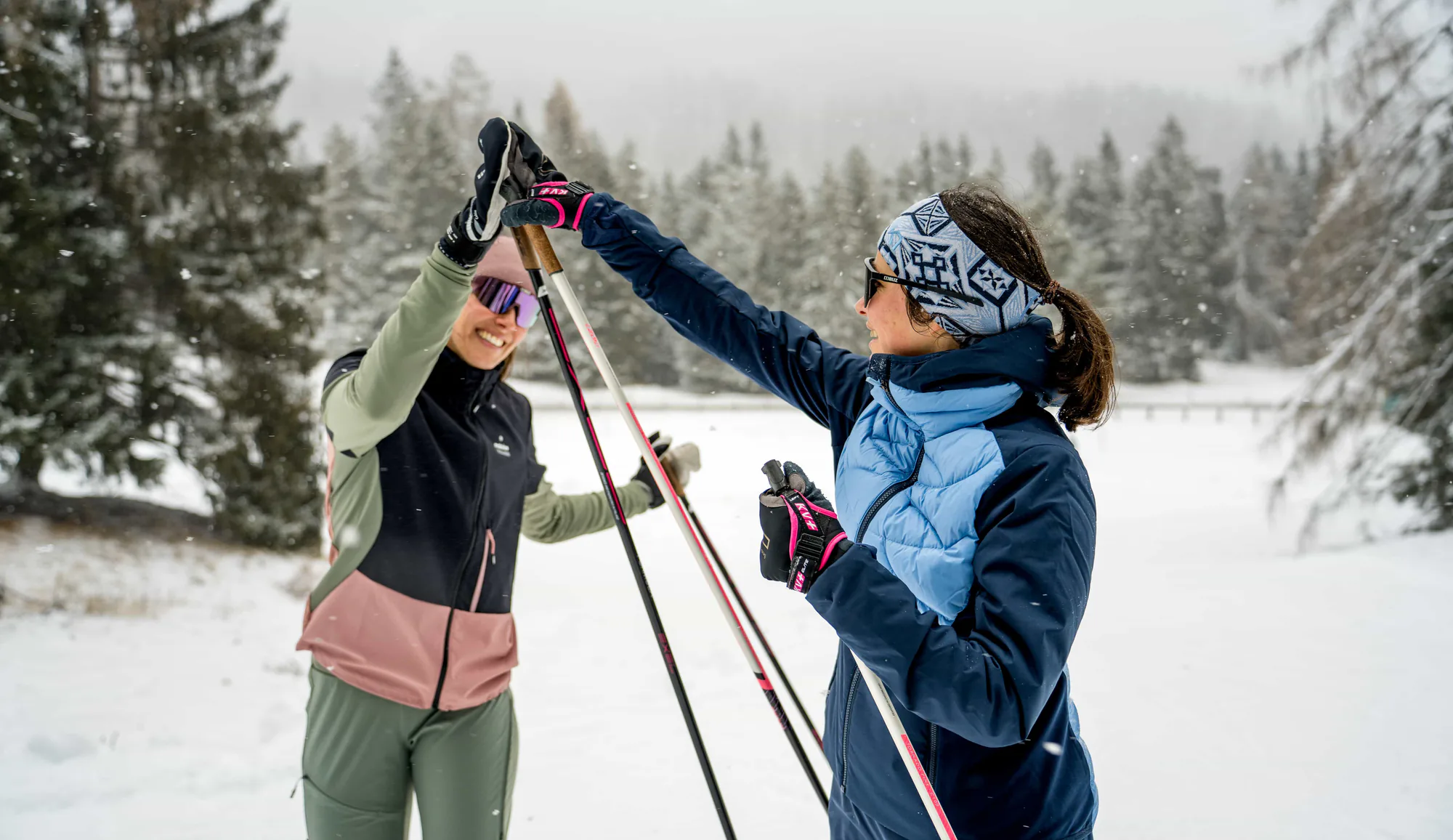 Zwei Langläuferinnen klatschen sich im Schneetreiben auf einer Loipe bei Lenzerheide ab, im Hintergrund verschneite Tannen.