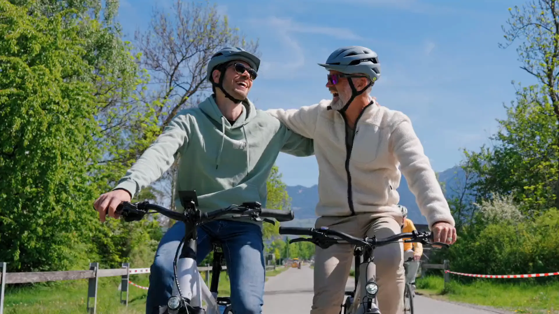 Zwei Radfahrer auf E-Bikes auf einem Radweg, der eine legt dem anderen im Fahren den Arm um die Schulter, beide lachen.