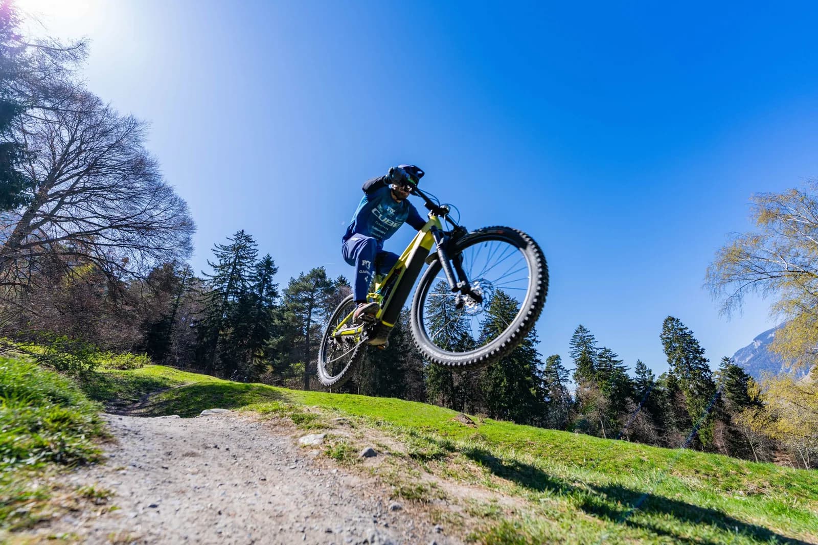 Aus der Froschperspektive: ein Fahrer springt mit einem gelben CUBE E-Mountainbike über eine Erdkante, beide Räder in der Luft vor tiefblauem Himmel.