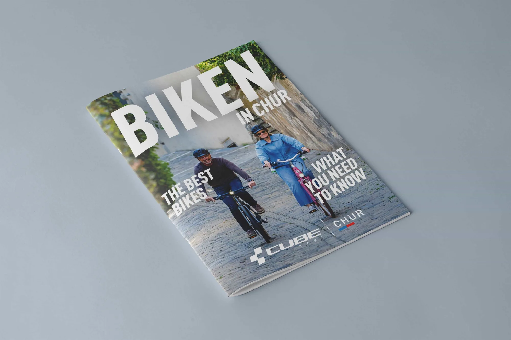 Das Magazin BIKEN IN CHUR liegt geschlossen auf hellgrauem Grund, auf dem Titel zwei Freizeitradler auf Kopfsteinpflaster und die Doppelmarke CUBE STORE Chur.