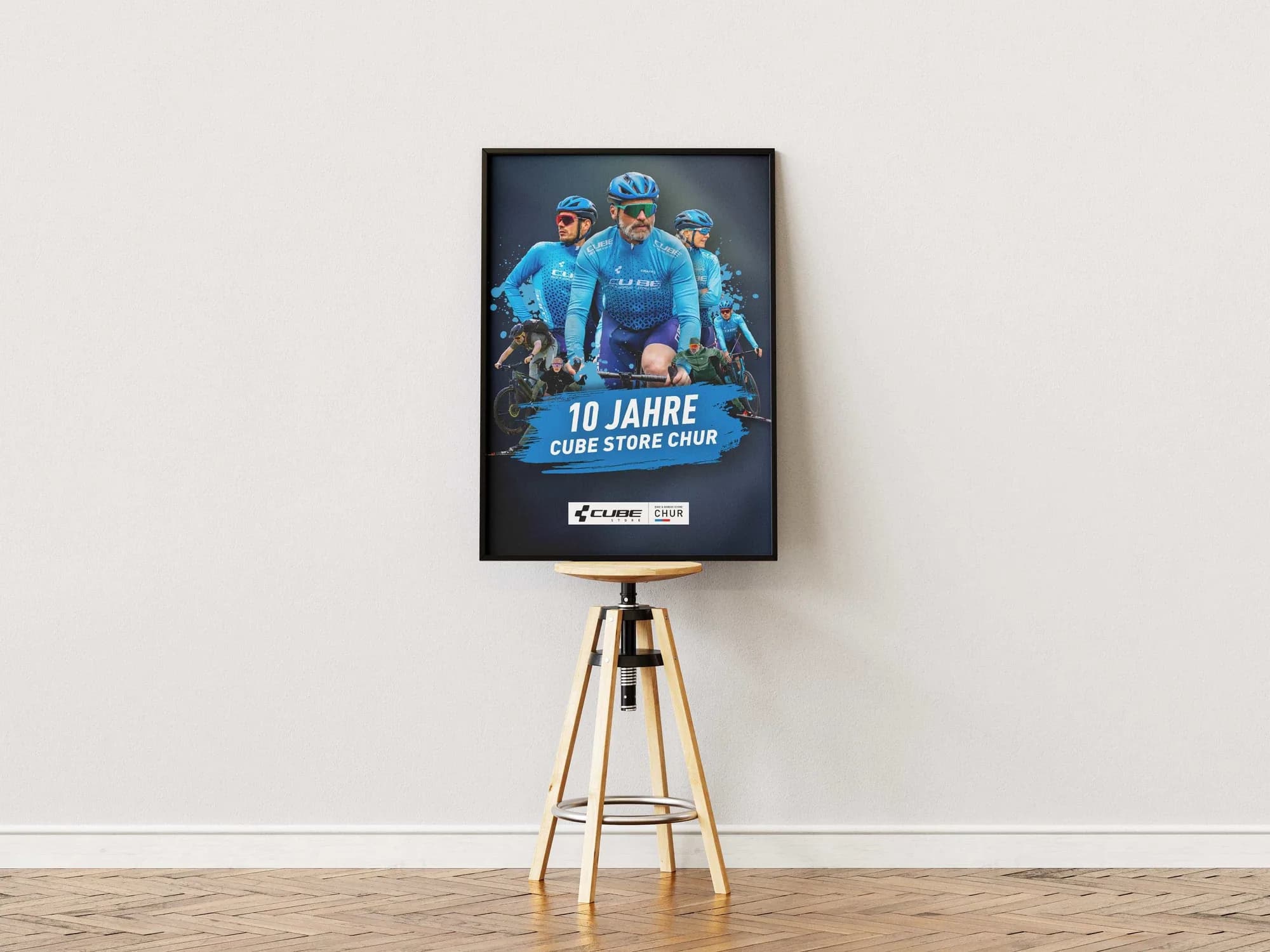 Ein schwarz gerahmtes Plakat auf einem hölzernen Dreibein-Hocker: freigestellte Fahrerinnen und Fahrer vor fast schwarzem Grund, die vordersten im blauen CUBE-Trikot, darunter der blaue Pinselstrich mit 10 JAHRE CUBE Store Chur.