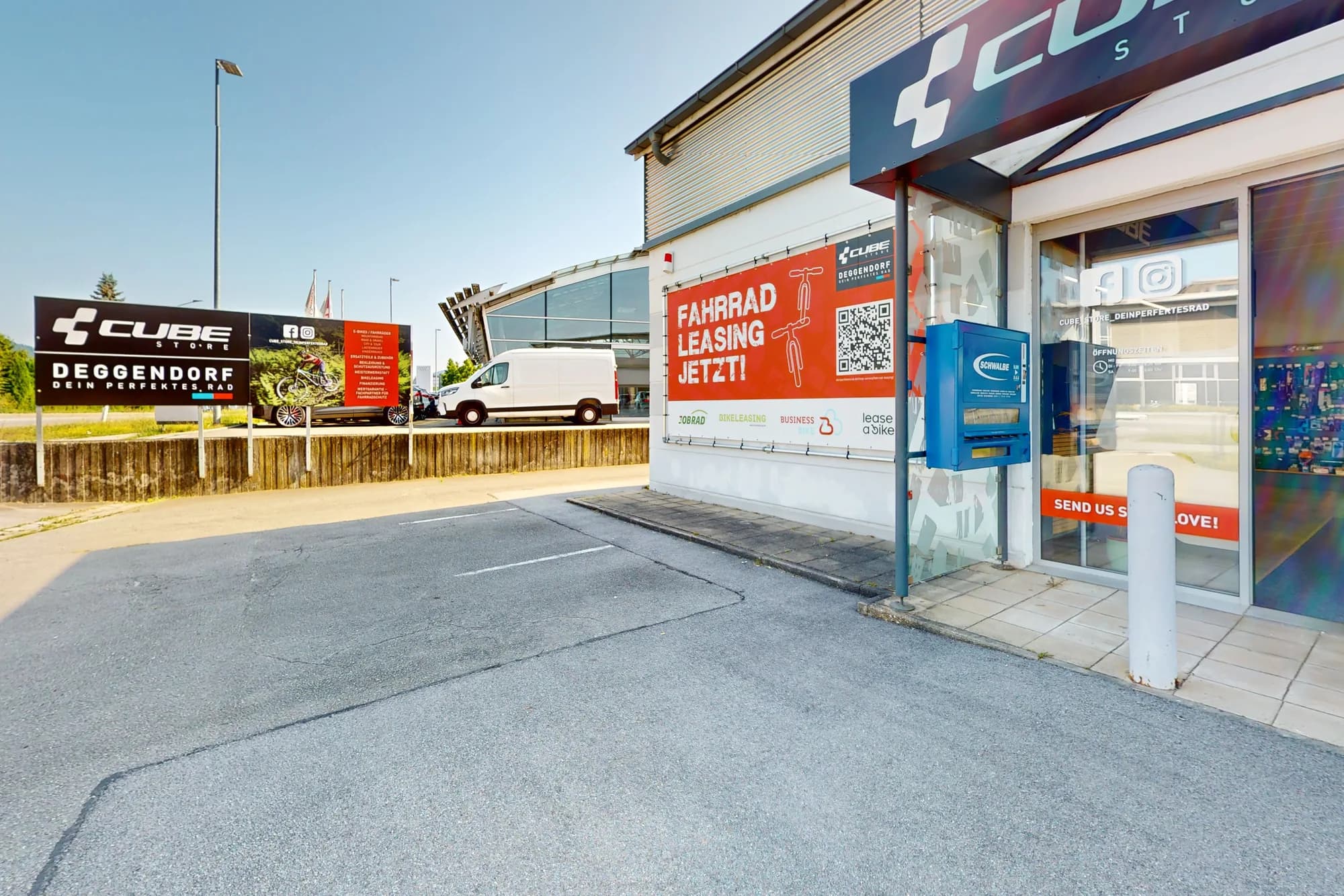 Die Ladenfront des CUBE Store Deggendorf vom Parkplatz aus: über der Fensterfront der blaue Fassadenstreifen mit dem CUBE-Zeichen, daneben das rote Plakat zum Fahrradleasing.