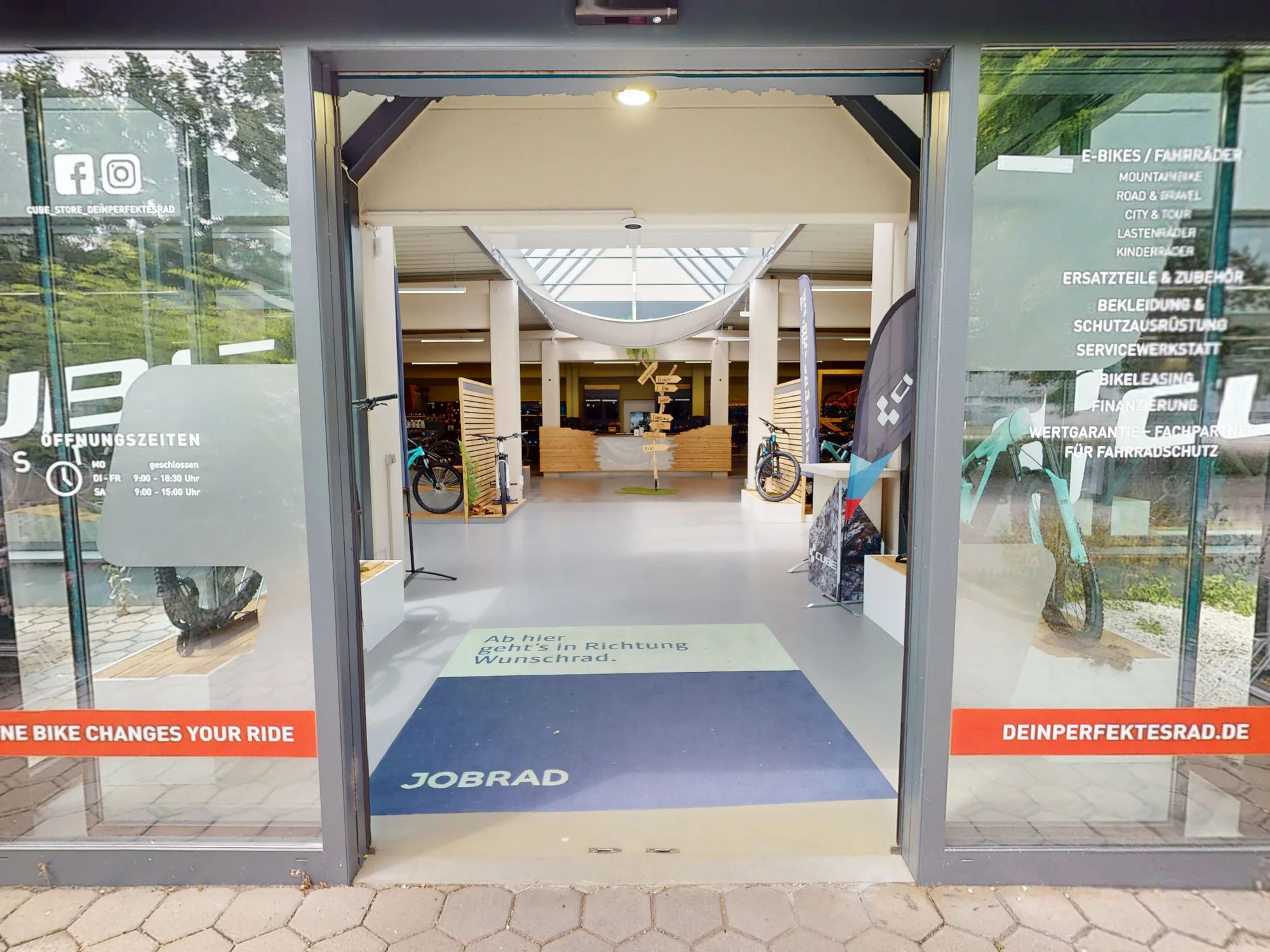 Der Eingang des CUBE Store Dingolfing von außen: eine breite Glastür mit den Aufklebern von JobRad und deinperfektesrad.de, dahinter der helle Verkaufsraum.