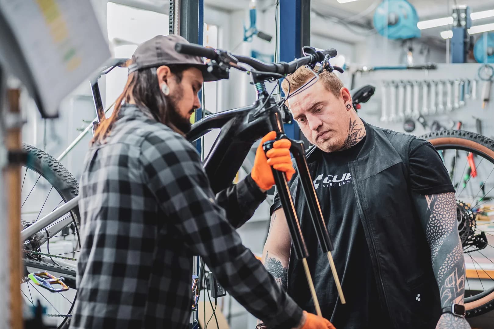 Zwei Mechaniker des CUBE Store Lindenberg arbeiten in der Werkstatt am selben Rad: einer hält die ausgebaute Federgabel in Arbeitshandschuhen, der andere das Mountainbike im Montageständer.