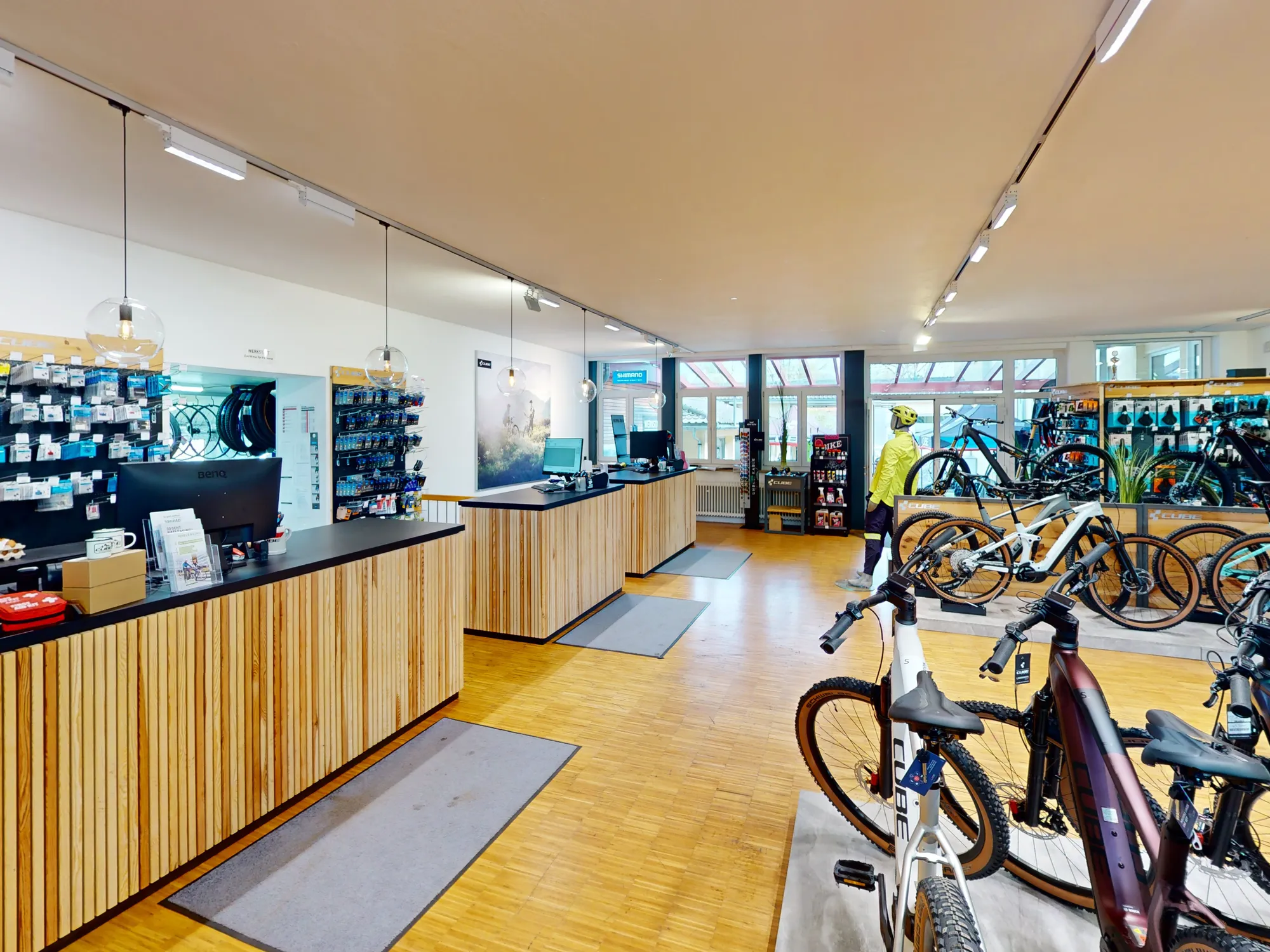 Die Theke im CUBE Store Lindenberg: eine lange Holzverkleidung mit zwei Beratungsplätzen und Bildschirmen, links die Zubehörwand, rechts die E-Bikes auf dem Podest.