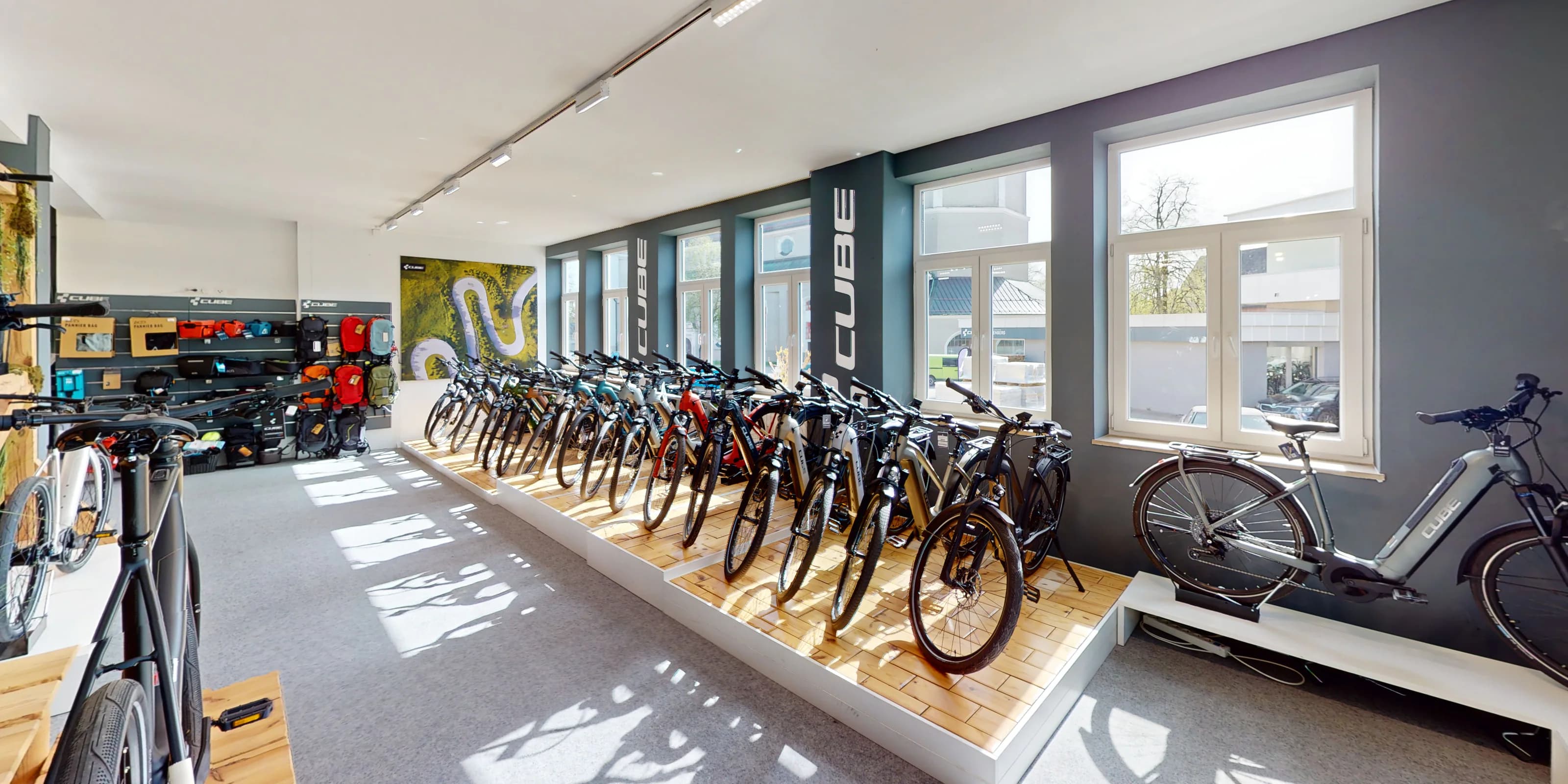 Virtueller Rundgang für den CUBE Store Lindenberg: Eine Reihe Trekkingräder auf einem Holzpodest an der Fensterwand des Verkaufsraums, darüber die CUBE-Schriftzüge auf den Pfeilern.