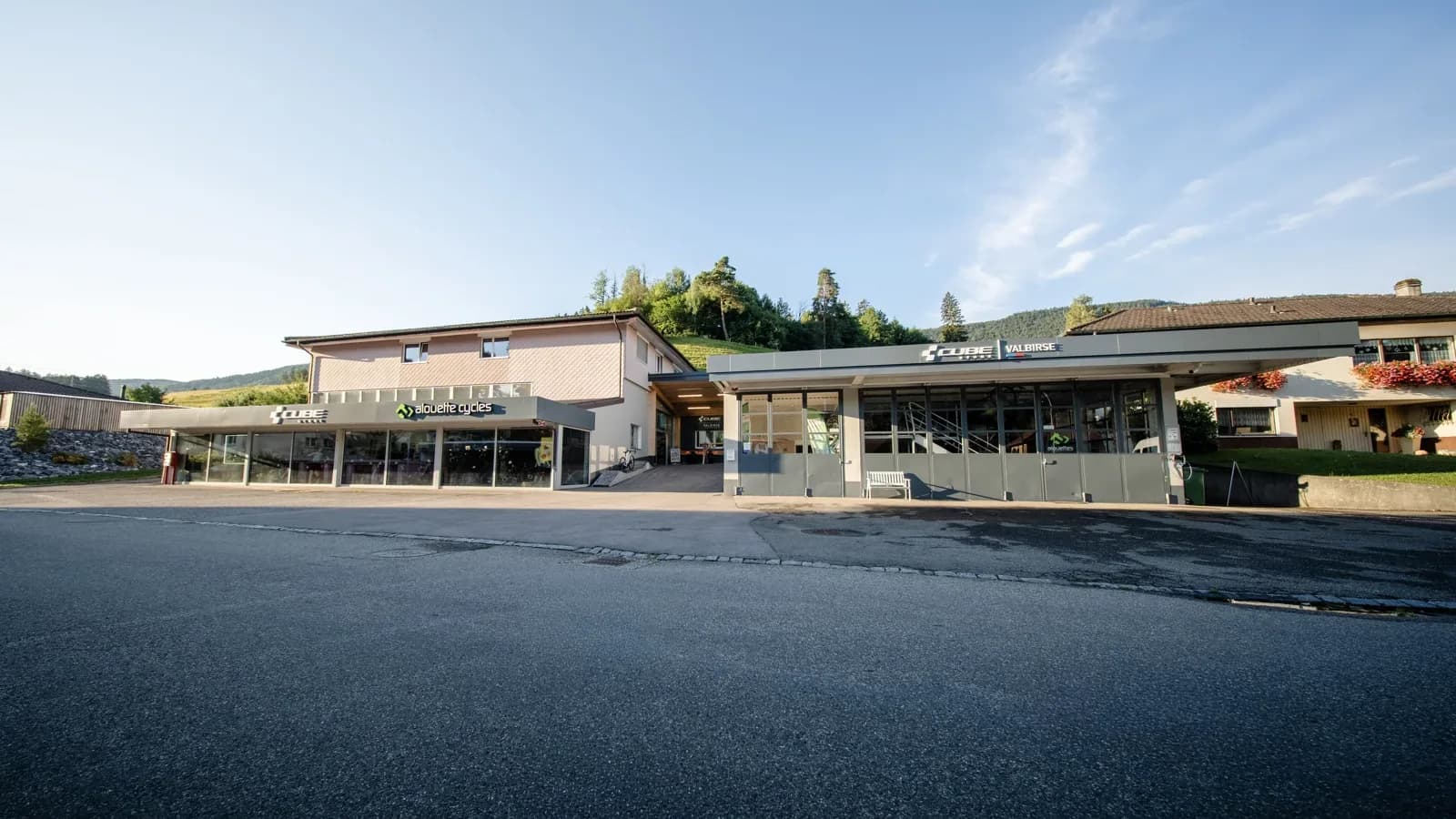 Der CUBE Store Valbirse vom Vorplatz aus: ein langgezogenes Gebäude mit den Schriftzügen alouette cycles und CUBE STORE VALBIRSE über den Schaufenstern.