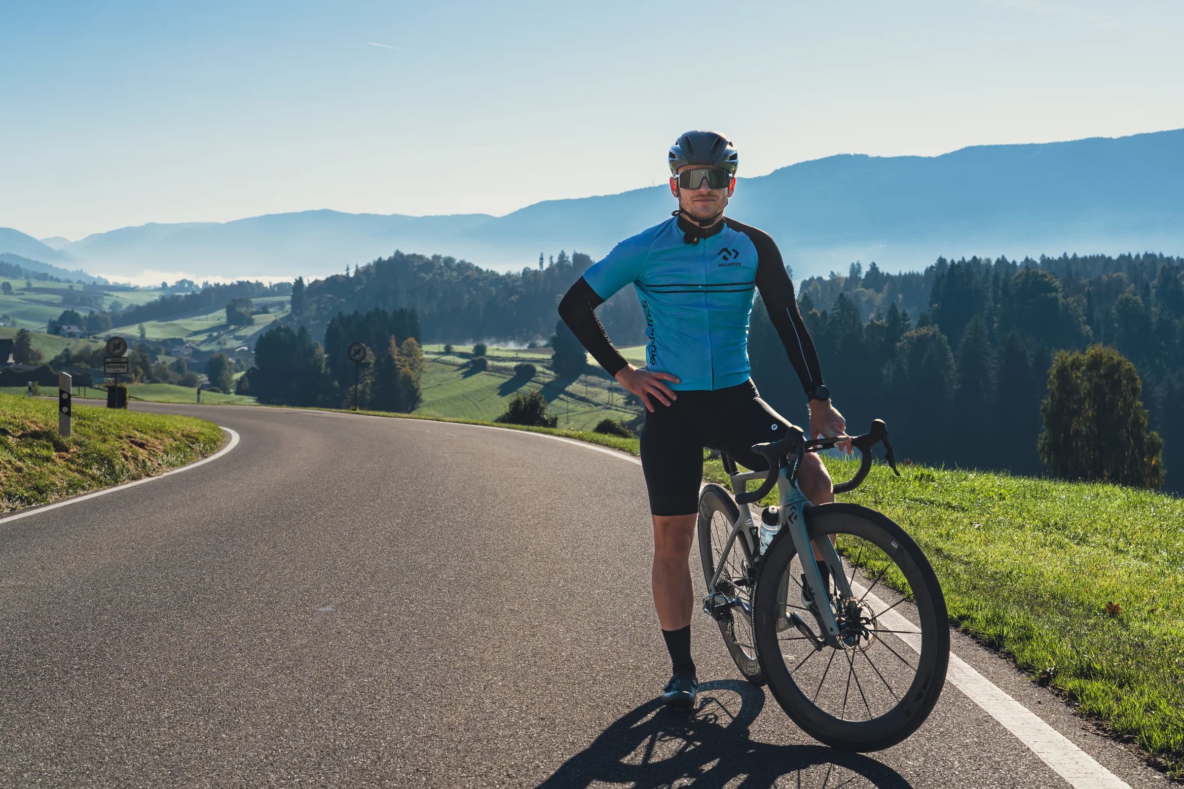 Eine Fahrerin steht mit ihrem Gravelbike auf einer Passstraße, die Hand in der Hüfte, dahinter Hügel und Himmel.