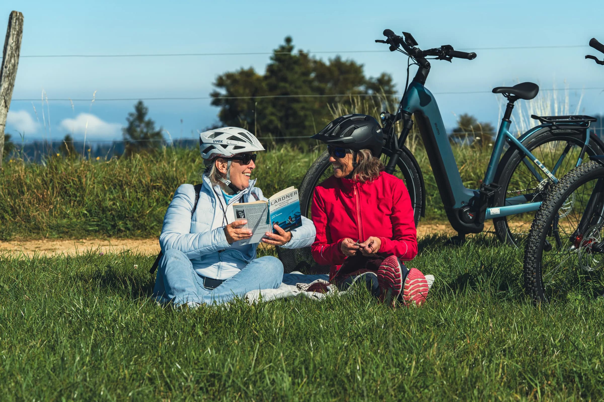 Zwei Frauen sitzen mit Helm im Gras und lesen, hinter ihnen lehnen zwei E-Bikes am Zaun.