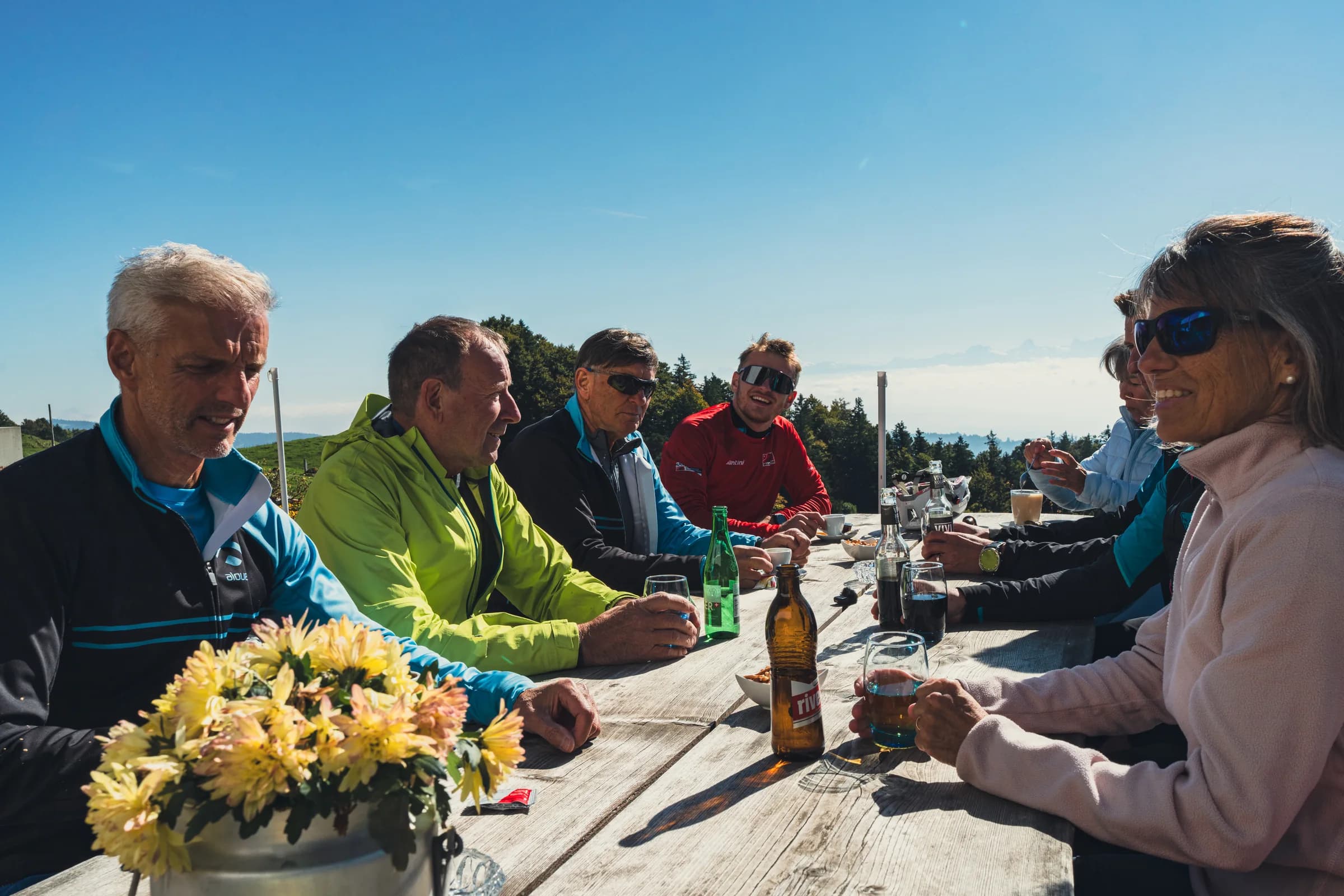 Eine Gruppe in Radkleidung sitzt an einem Tisch im Freien und stößt mit Gläsern an, Blumen auf dem Tisch.