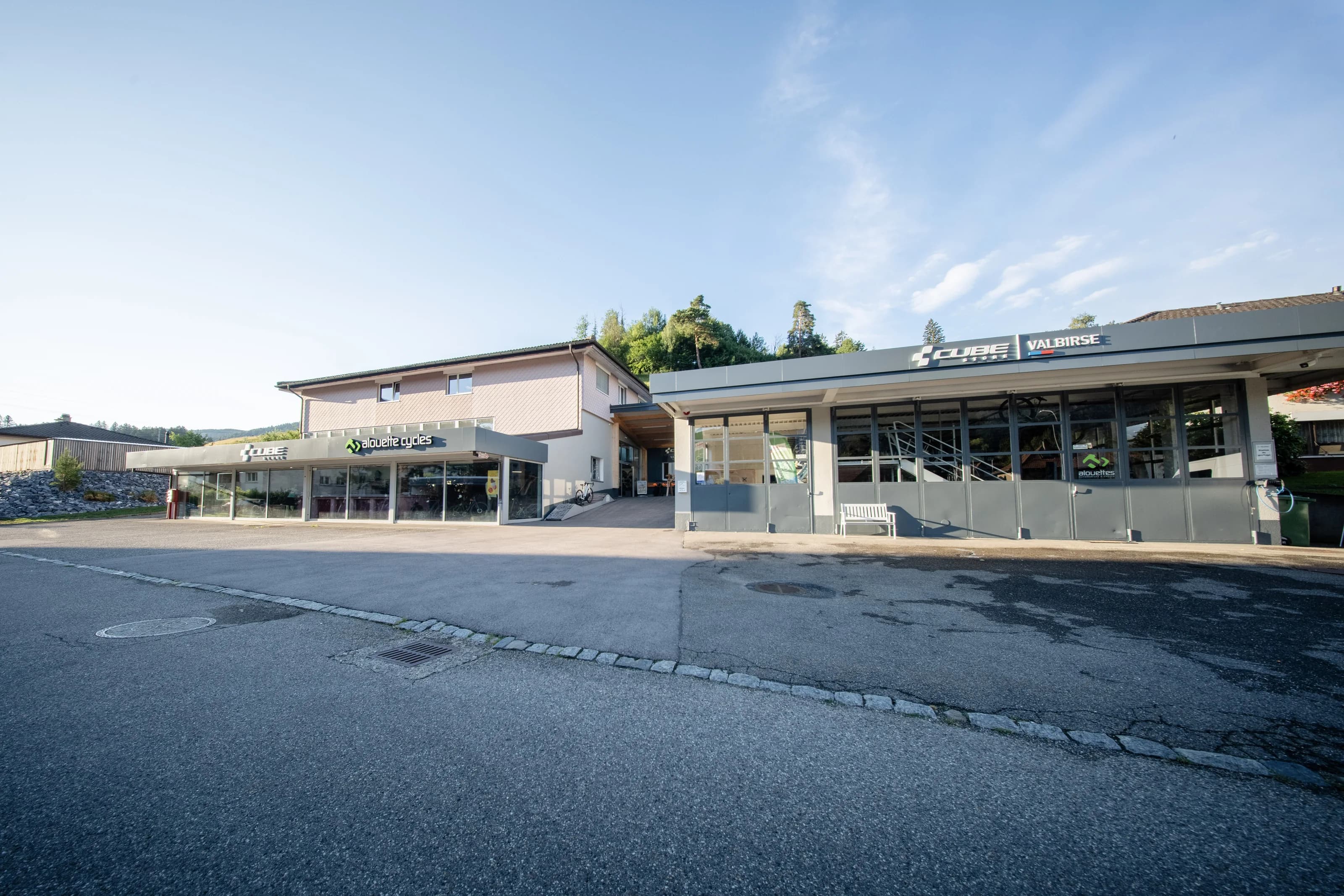 CUBE Store Valbirse: Die Ladenfront mit der Leuchtschrift CUBE Store Valbirse über den Schaufenstern, davor der Vorplatz und dahinter der bewaldete Jura.