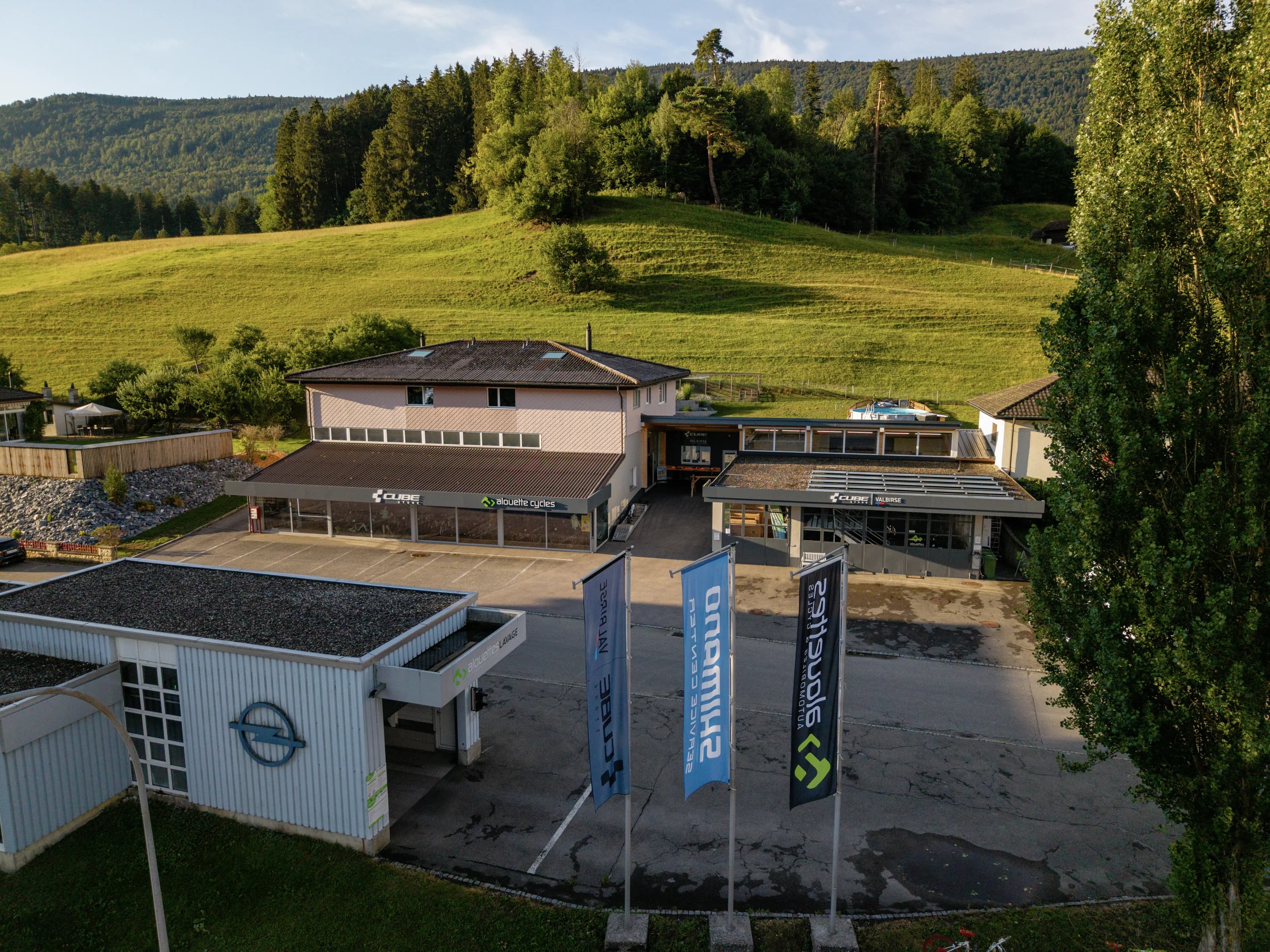 Der CUBE Store Valbirse aus der Luft: das Gebäude mit den Fahnen von CUBE und Shimano am Vorplatz, dahinter die Wiese und der Waldrand.