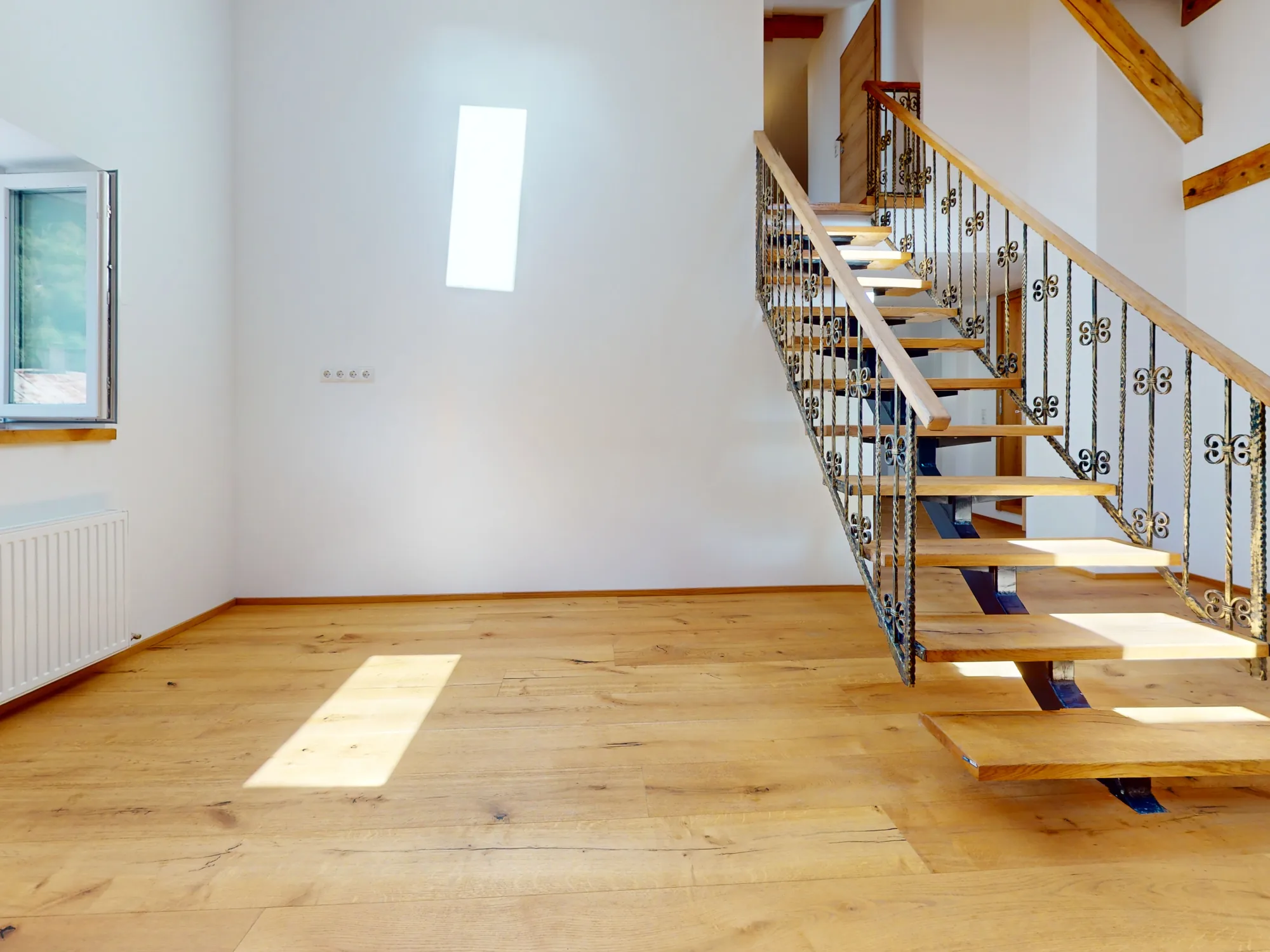Ein sanierter Wohnraum in der Maisengasse: helle Eichendielen mit einem Sonnenfeld darauf, rechts eine schmiedeeiserne Treppe ins Obergeschoss, darüber ein sichtbarer Holzbalken.