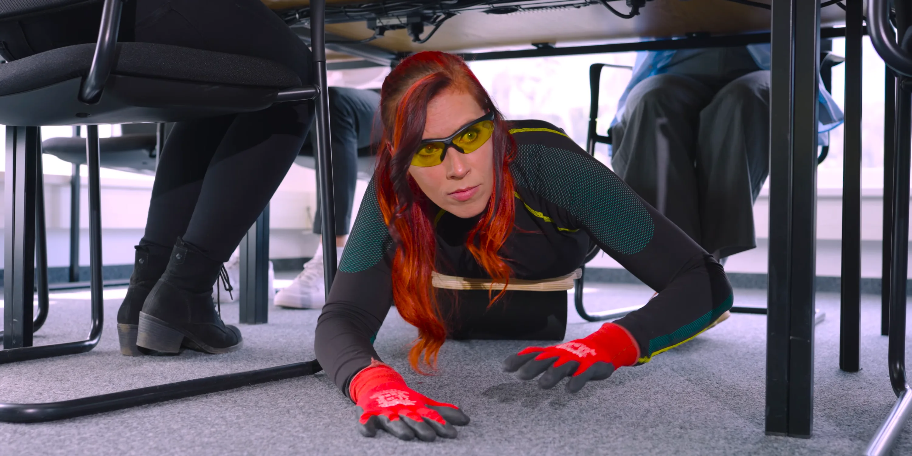 Eine Frau mit roten Haaren, gelber Schutzbrille und roten Arbeitshandschuhen robbt unter einem Bürotisch hervor, Standbild aus der Agentenfolge der Erklärvideo-Serie für Exceedation Sales.