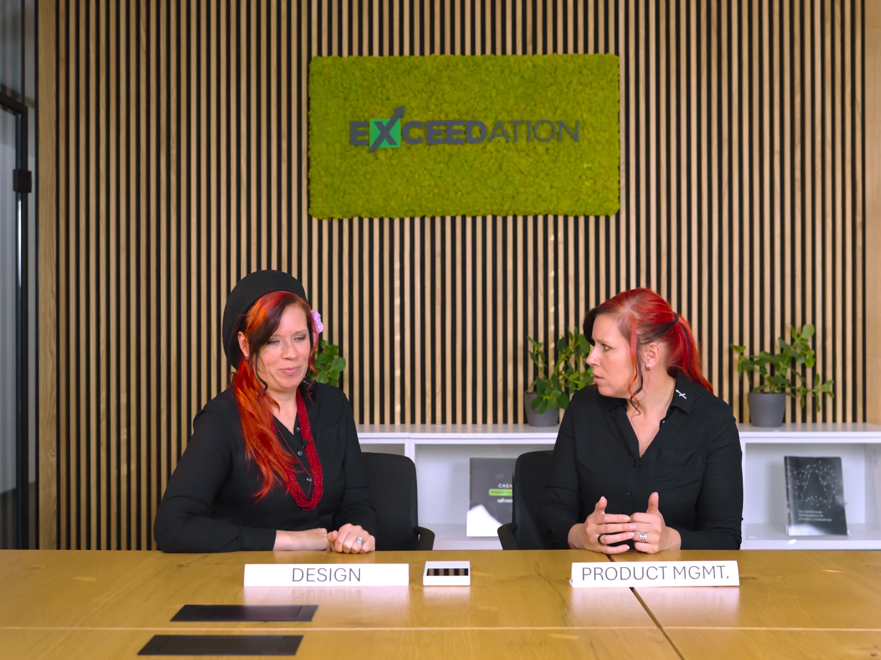 Zwei Frauen mit roten Haaren sitzen an einem Besprechungstisch vor den Schildern Design und Product Mgmt., dahinter eine Holzlamellenwand mit dem Exceedation-Logo aus Moos, Standbild aus Folge 9 der Videoserie.