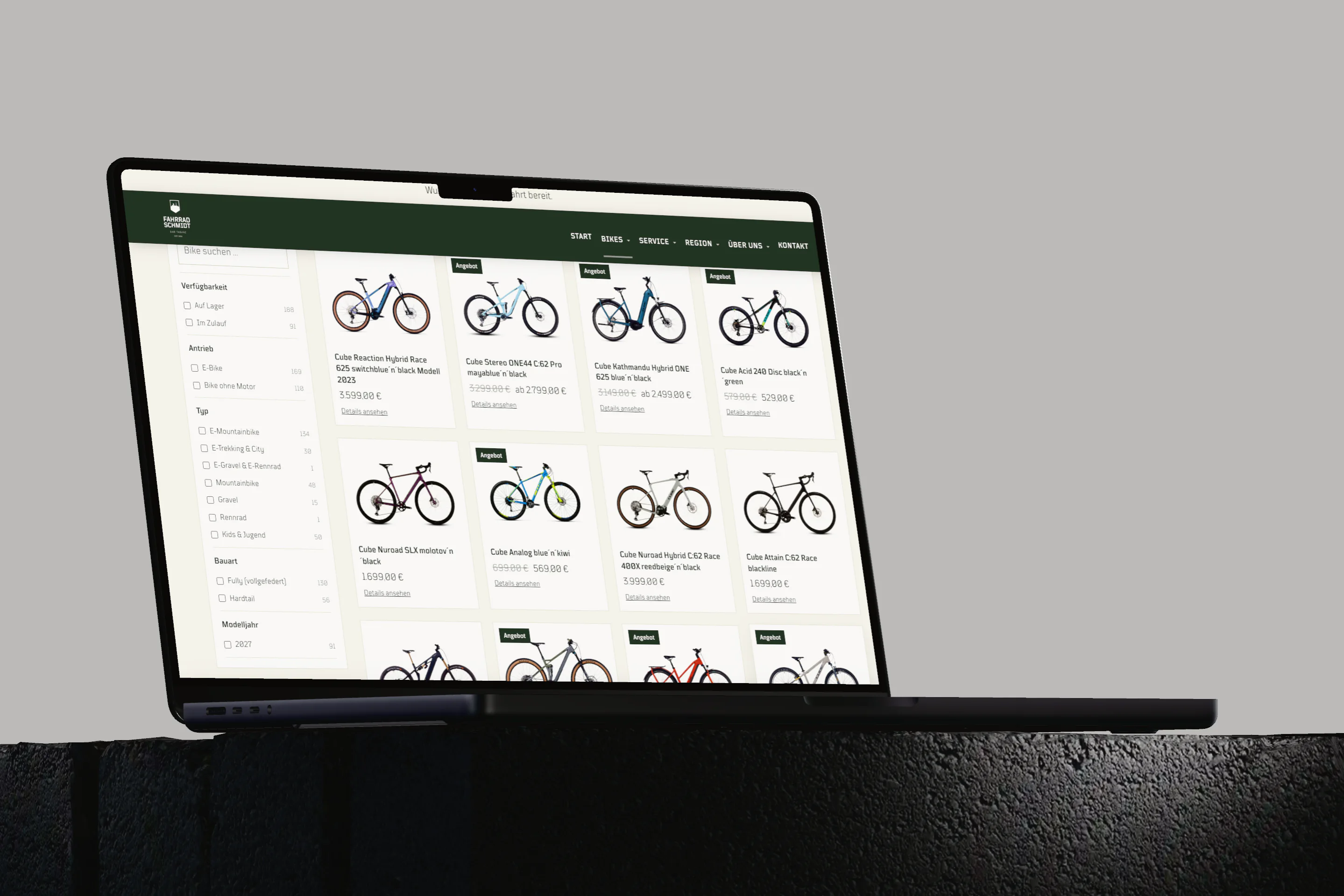 Ein MacBook Pro auf dunklem Sockel zeigt den Showroom von fahrrad-schmidt.de: links die Filterspalte, rechts das Raster der Räder mit Preisen.