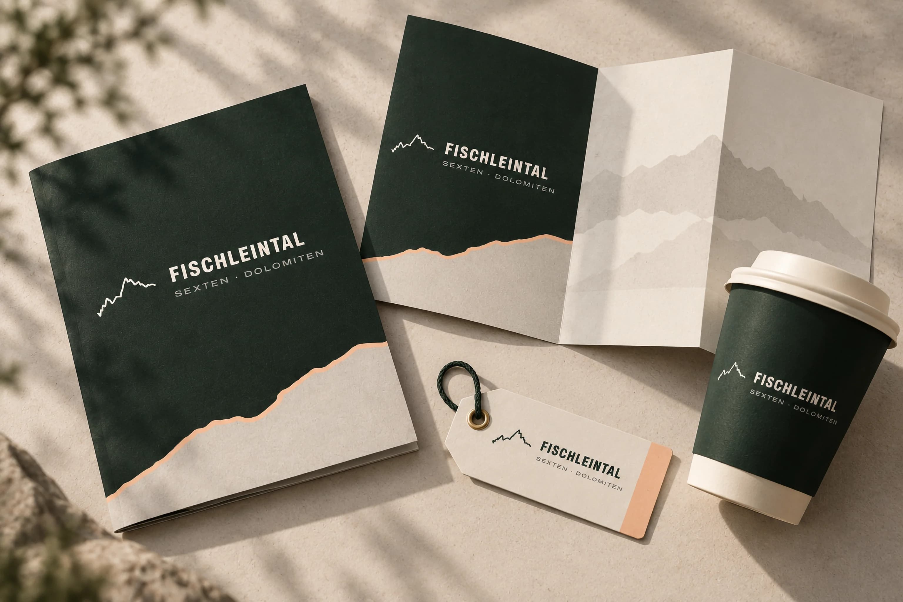 Eine waldgrüne Mappe und ein aufgeklappter Flyer mit Bergsilhouette in Grau, dazu ein heller Anhänger mit dem Logo des Fischleintals und ein waldgrüner Becher auf hellem Stein.