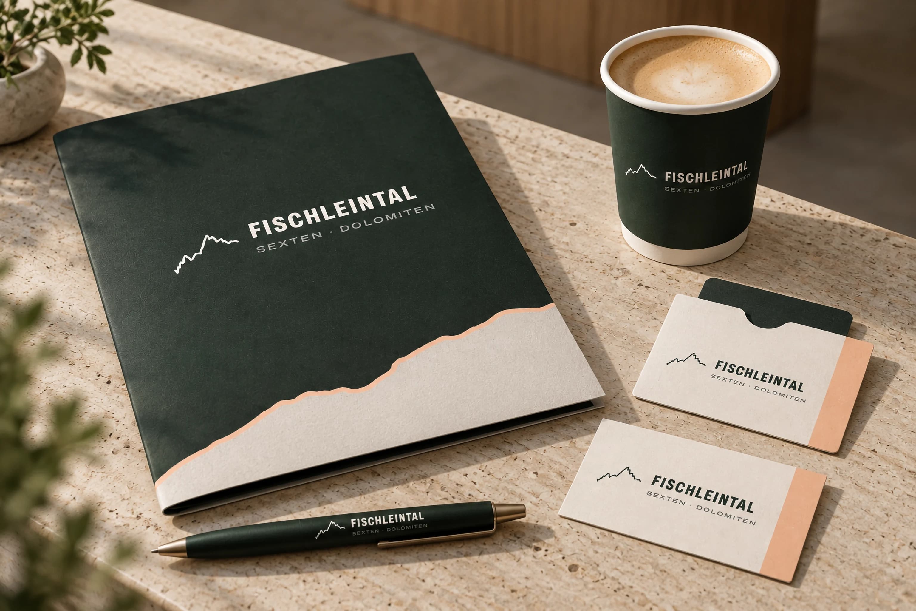 Eine waldgrüne Mappe mit dem Logo des Fischleintals und einer hellen Bergkante am unteren Rand, daneben ein Becher, zwei helle Karten mit Logo und Pfirsichstreifen und ein grüner Stift auf einem Steintisch.