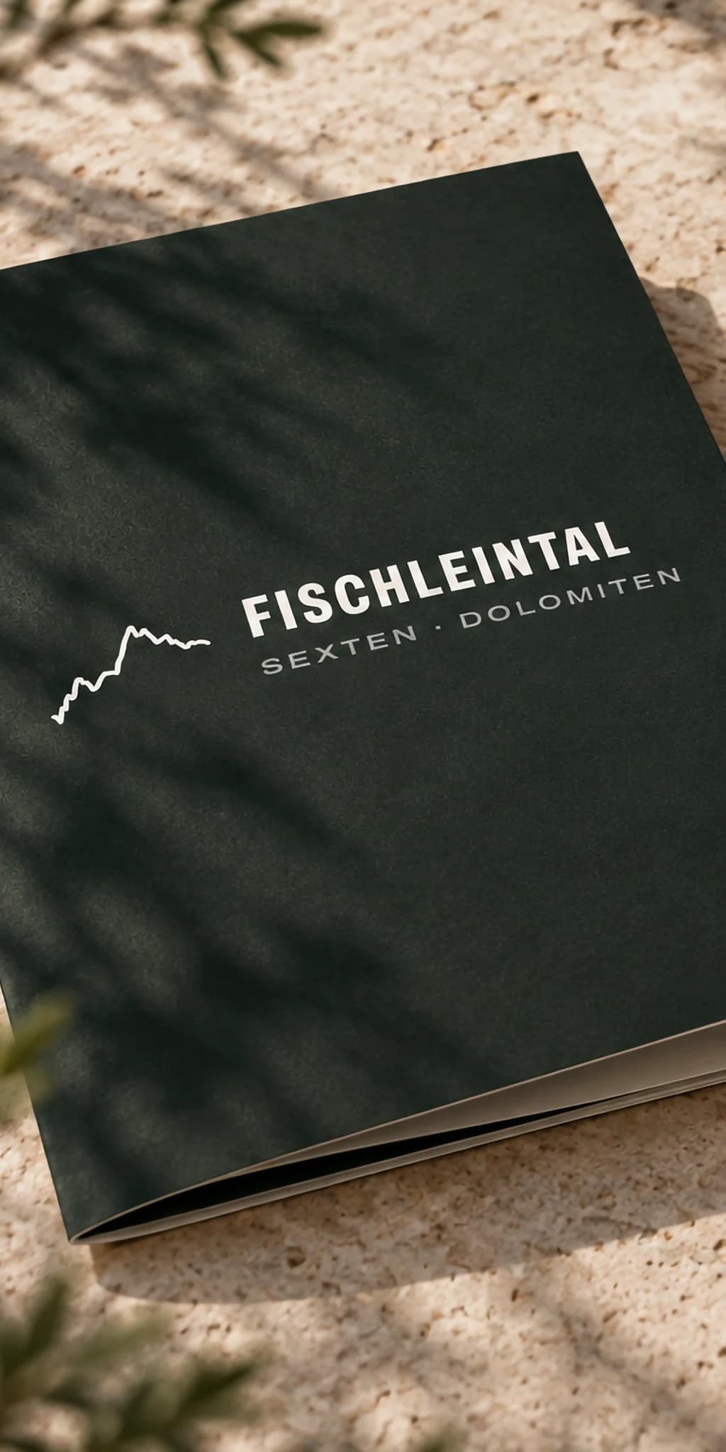 Geschäftsausstattung des Fischleintals auf einem hellen Steintisch im Sonnenlicht: eine waldgrüne Mappe mit dem Logo in Weiß, ein Becher in Waldgrün, ein helles Notizbuch und eine helle Karte mit Logo und einem Streifen in Pfirsich.
