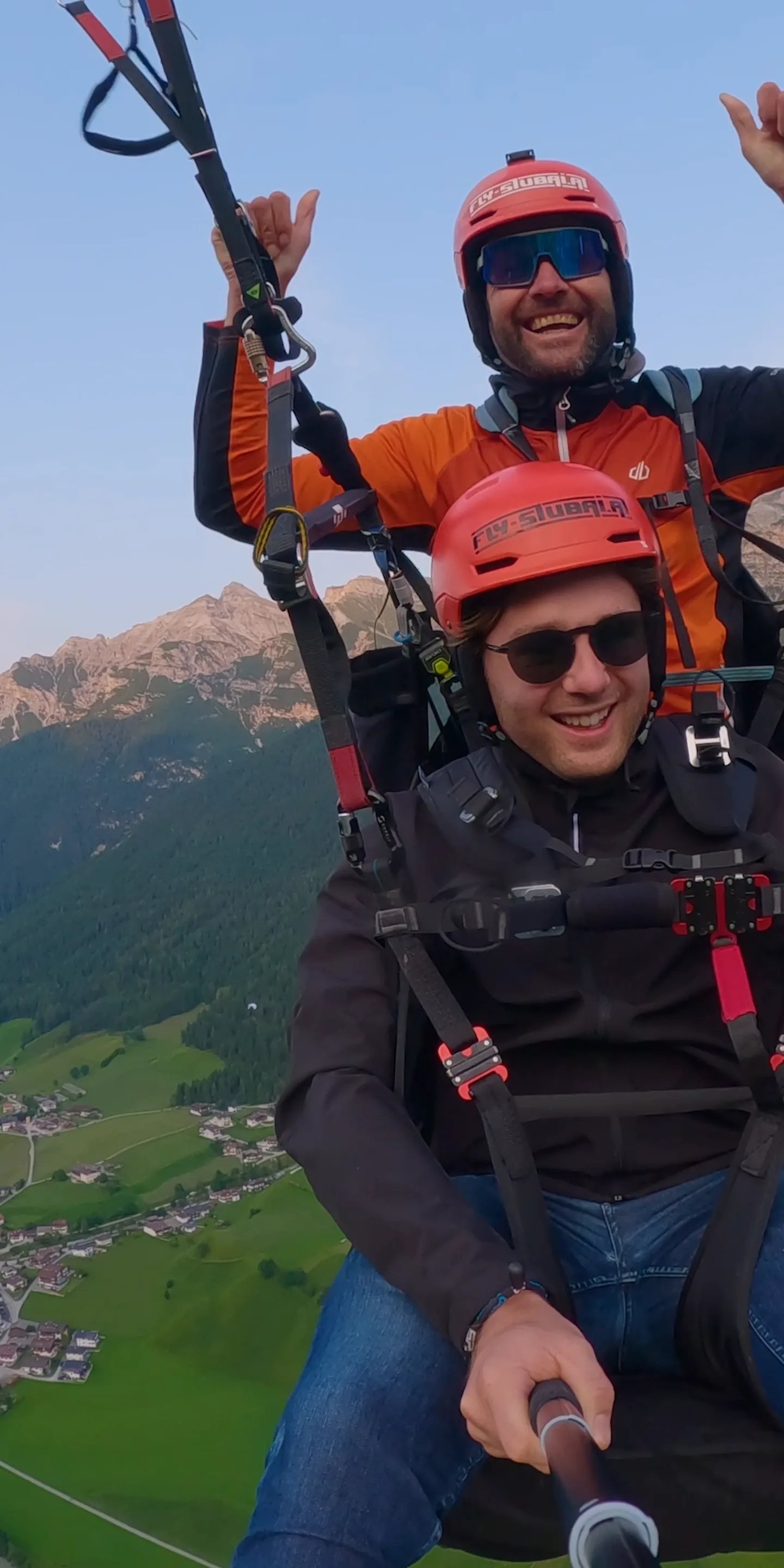 Pilot und Gast hängen im Tandem-Gurtzeug hoch über dem grünen Stubaital, beide mit roten Helmen, dahinter die Berge im Abendlicht.