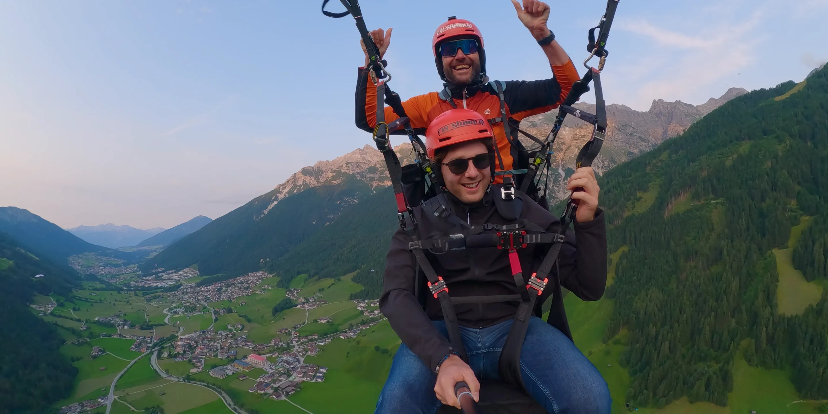 Fly Stubai: Pilot und Gast hängen im Tandem-Gurtzeug hoch über dem grünen Stubaital, beide mit roten Helmen, dahinter die Berge im Abendlicht.