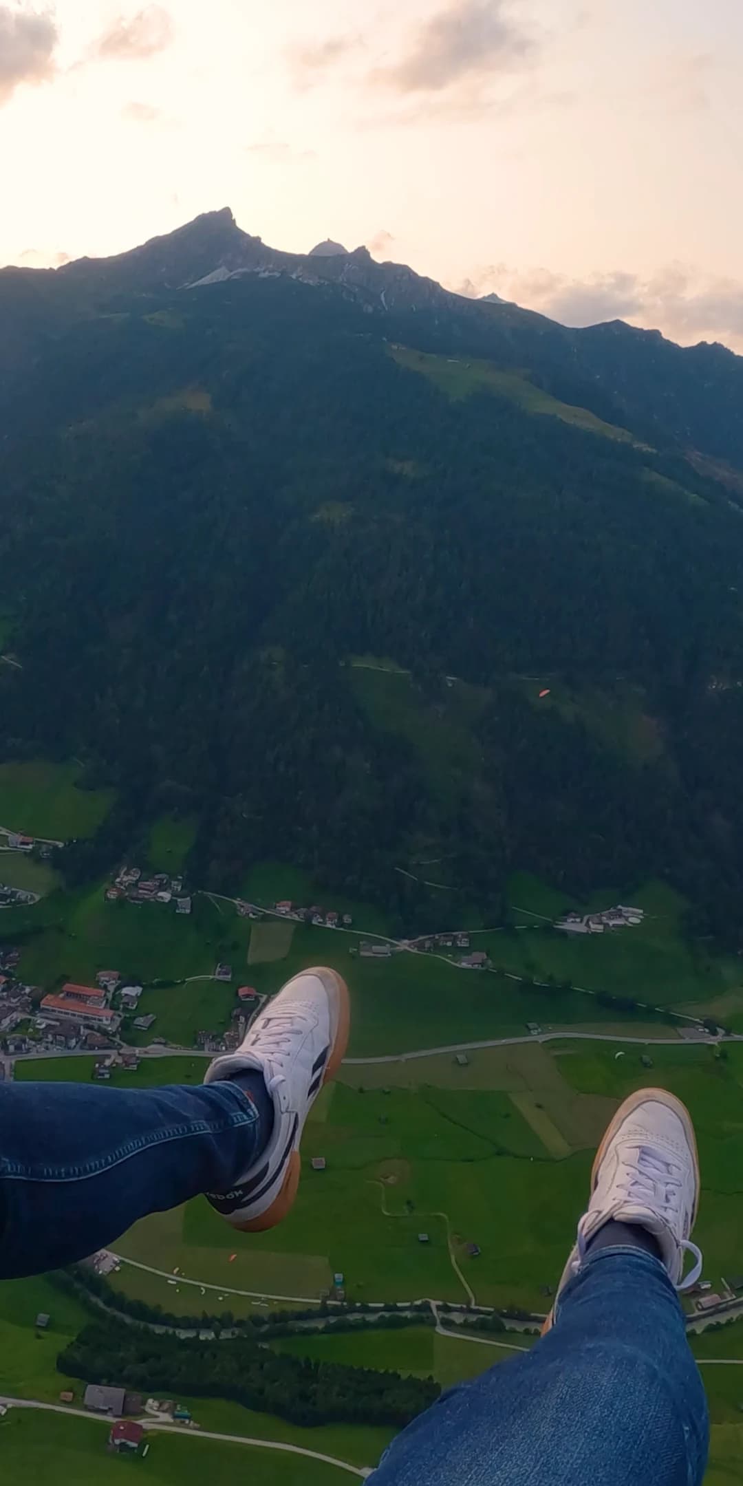 Blick nach unten aus dem Tandem im Abendlicht: die eigenen Beine in Jeans und weißen Turnschuhen über dem Stubaital mit seinen Dörfern und Wiesen, dahinter die Berge unter einem hellen Abendhimmel.