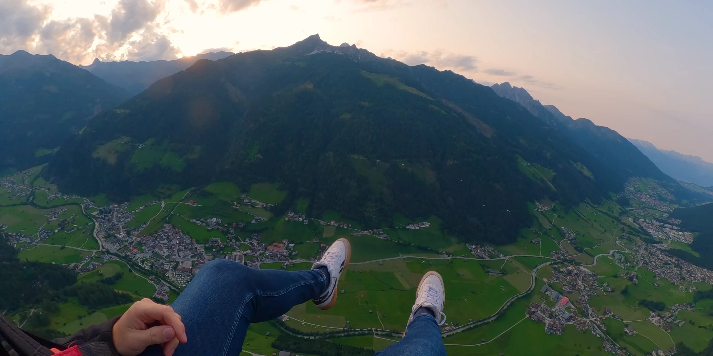Blick nach unten aus dem Tandem im Abendlicht: die eigenen Beine in Jeans und weißen Turnschuhen über dem Stubaital mit seinen Dörfern und Wiesen, dahinter die Berge unter einem hellen Abendhimmel.