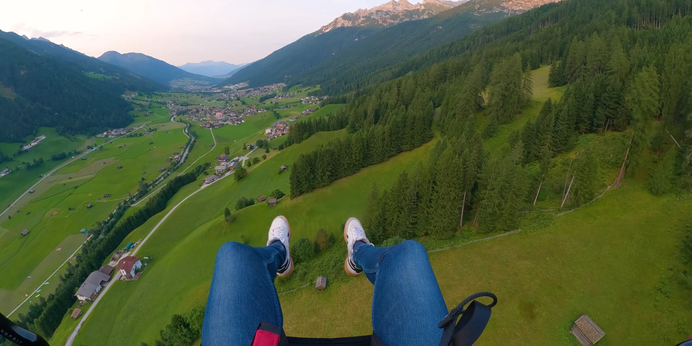 Blick nach unten aus dem Tandem: die eigenen Beine und Turnschuhe über dem grünen Stubaital mit Wiesen, Wald und den Häusern von Neustift.