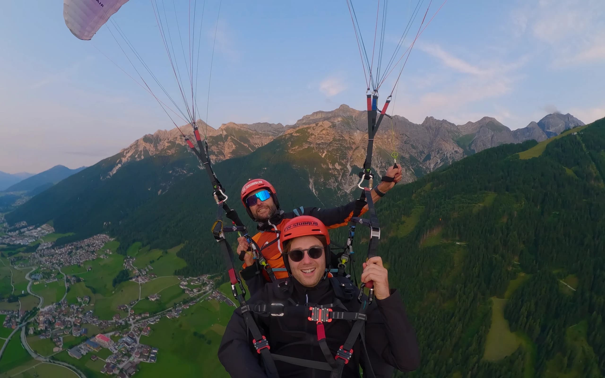 Pilot und Gast im Tandem über dem Stubaital im Abendlicht, vorne lacht der Gast mit rotem Helm und Sonnenbrille in die Kamera, dahinter die Gipfel im letzten Licht und das Tal mit dem Ort.