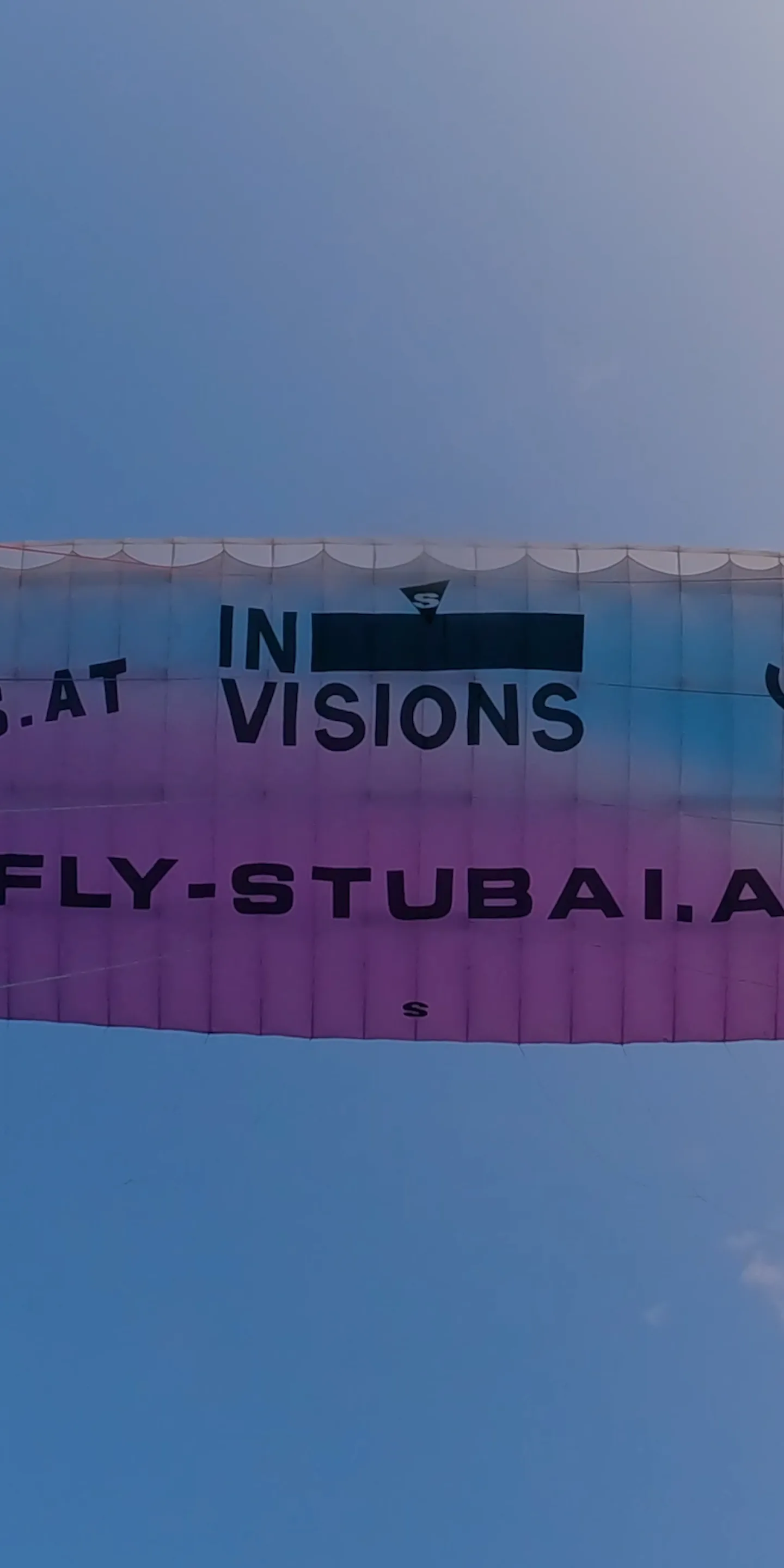 Der Gleitschirm von unten vor blauem Himmel, auf der Kappe die Schriftzüge Invisions und fly-stubai.at, die Leinen laufen zum Rand des Bildes.