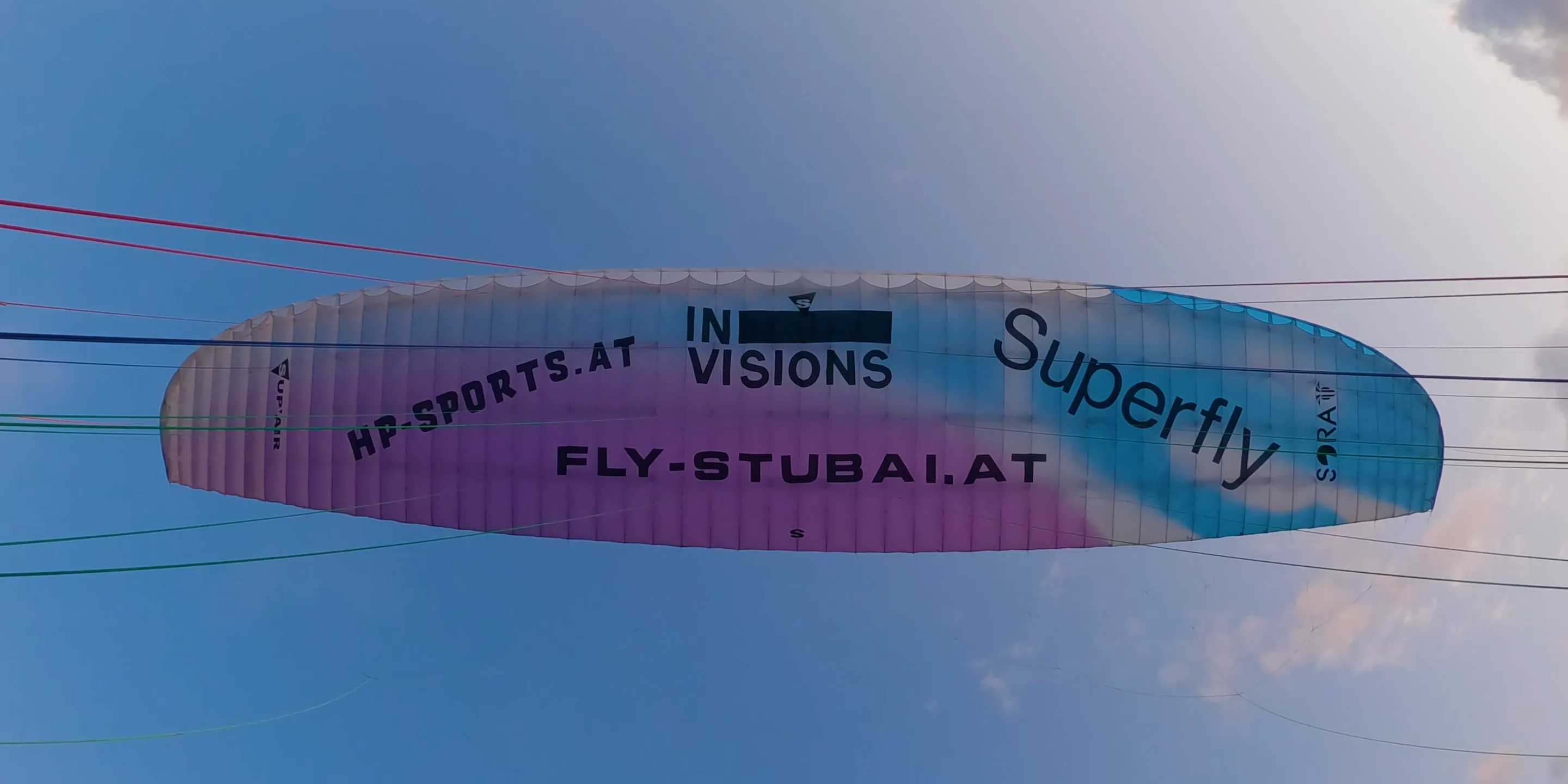 Der Gleitschirm von unten vor blauem Himmel, auf der Kappe die Schriftzüge Invisions und fly-stubai.at, die Leinen laufen zum Rand des Bildes.