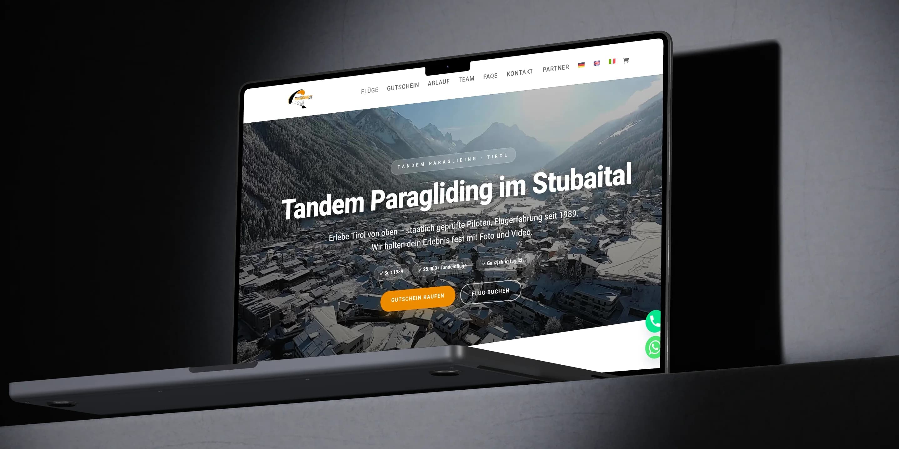 Ein MacBook im dunklen Studio zeigt die Startseite von fly-stubai.at: der verschneite Ort im Stubaital von oben, darüber die Überschrift Tandem Paragliding im Stubaital und die Knöpfe Gutschein kaufen und Flug buchen.