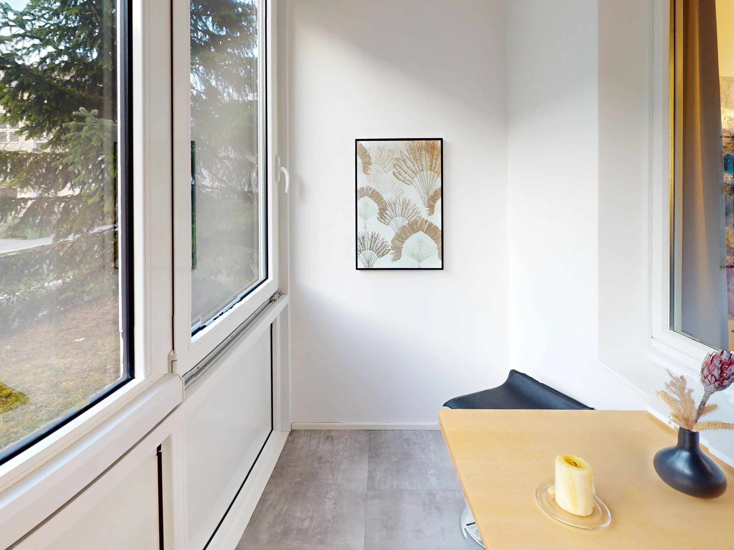 Eine Schreibtischecke am Fenster der eingerichteten Wohnung: heller Tisch mit Vase und Kerze, daneben eine bodentiefe Fensterfront mit Blick auf Bäume und Häuser.