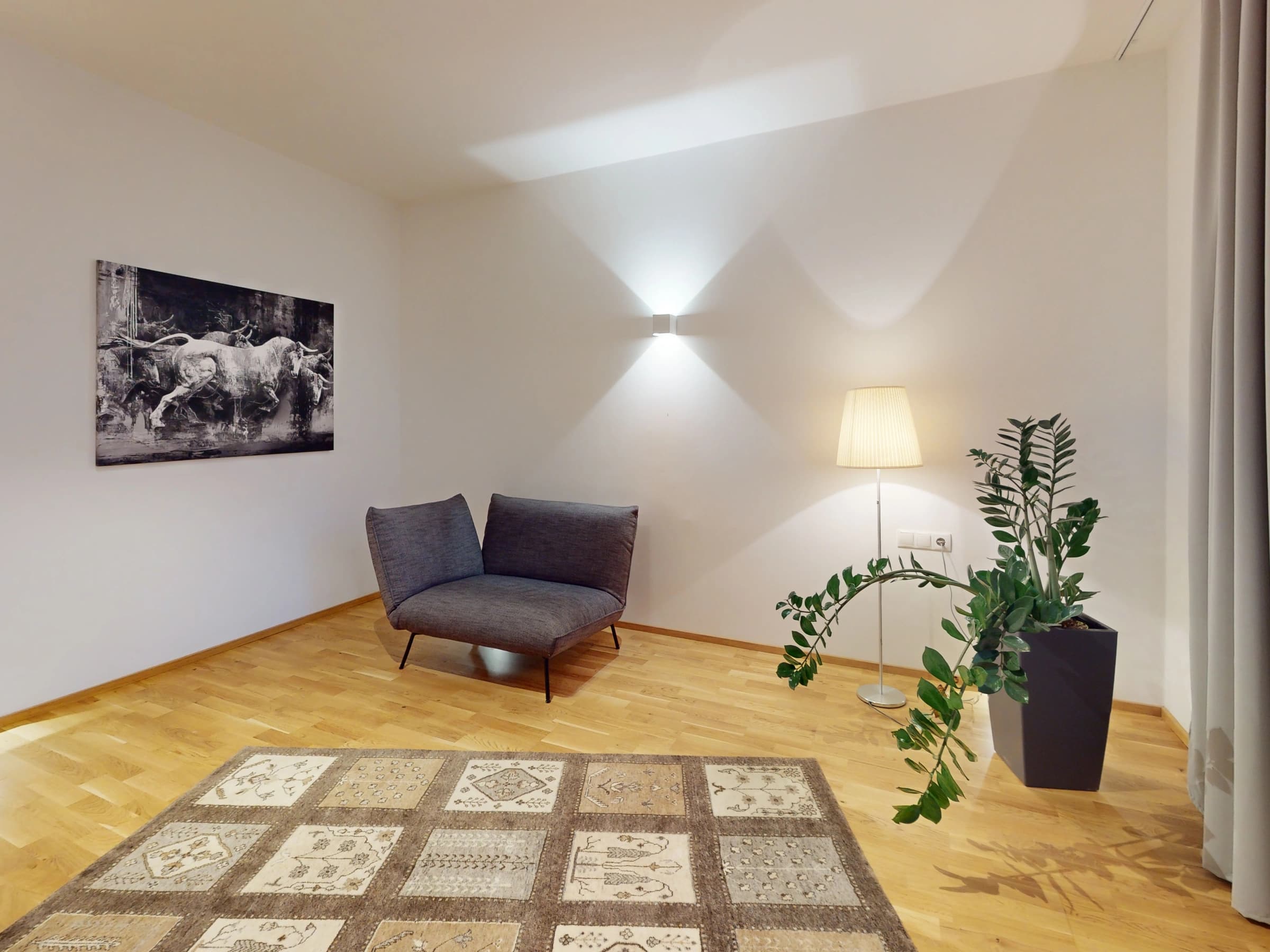 Wohnraum der eingerichteten Wohnung: eine graue Chaiselongue vor einer weißen Wand mit einem großformatigen Pferdefoto, daneben eine Stehlampe und zwei Grünpflanzen in dunklen Töpfen, davor ein gemusterter Teppich auf Parkett.