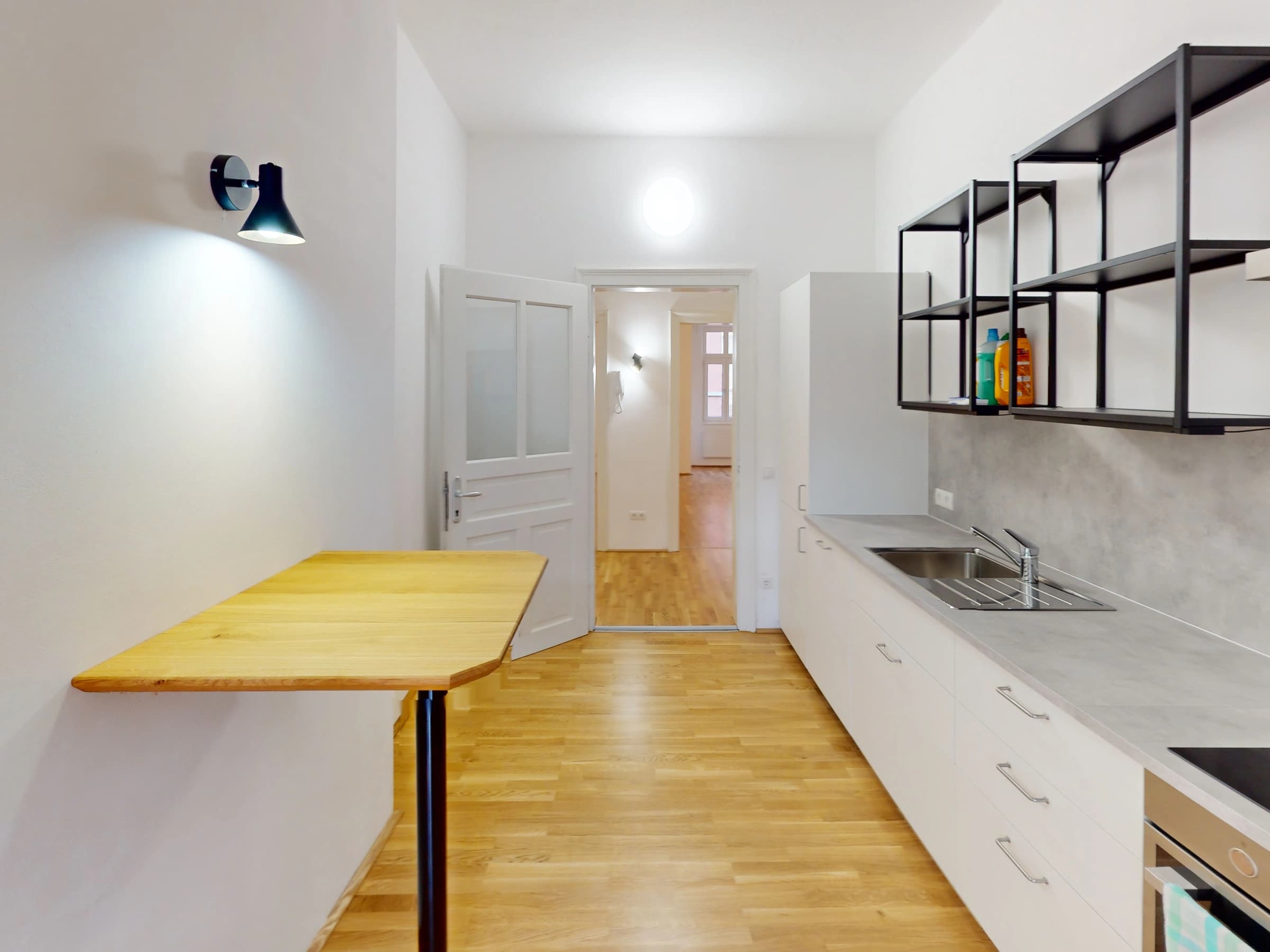 Die Küche der leeren Wohnung: links ein schmaler Holztisch auf schwarzem Fuß, in der Mitte eine offene Tür in den nächsten Raum, rechts die weiße Küchenzeile mit Spüle unter einem offenen schwarzen Stahlregal.