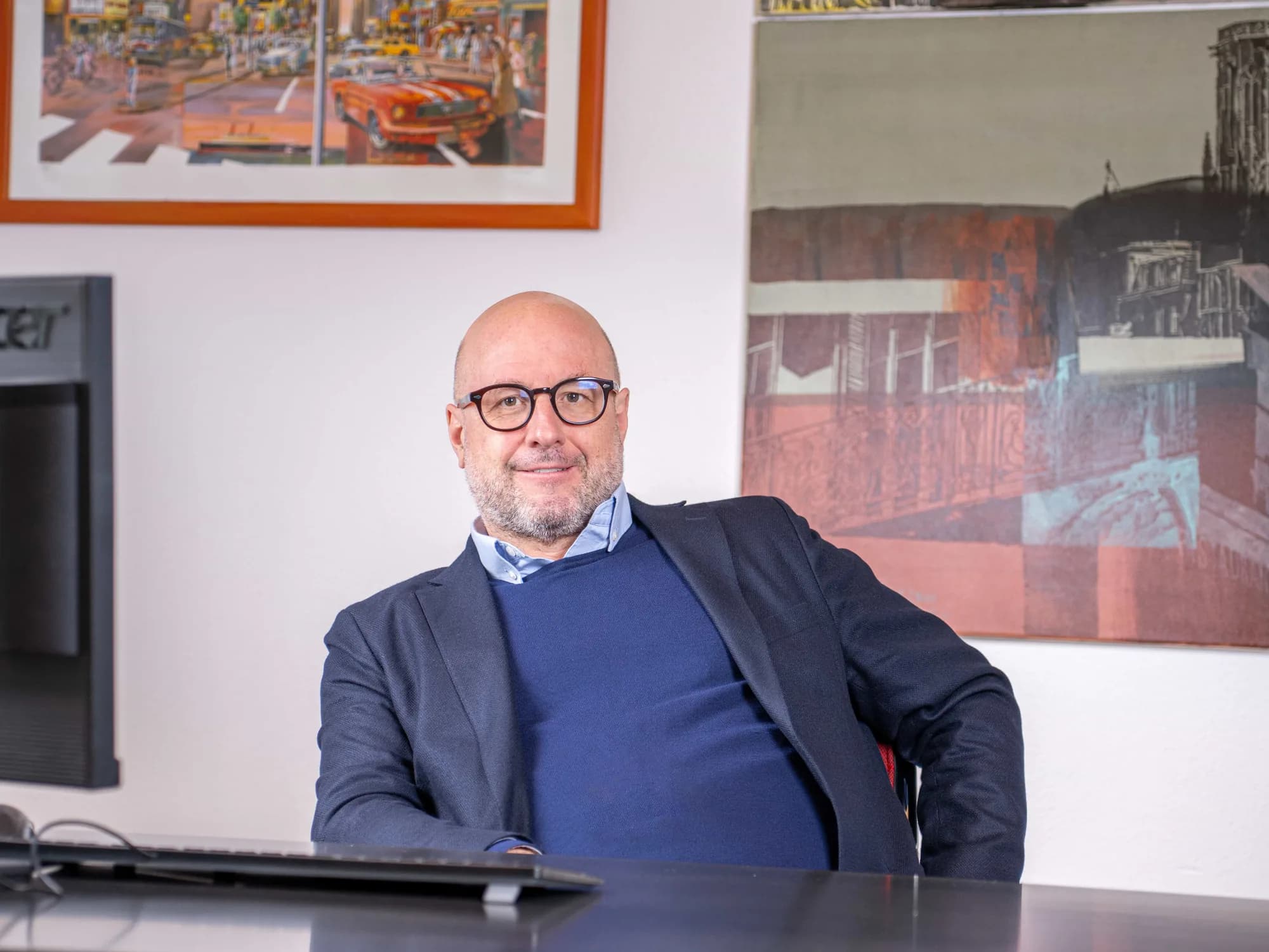 Ein Anwalt sitzt entspannt an seinem Schreibtisch und blickt in die Kamera, hinter ihm eine Wand mit gerahmten farbigen Bildern, links ein Bildschirm.