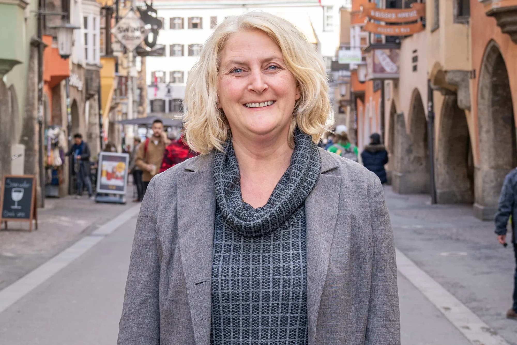 Einzelporträt in der Altstadt: eine Frau im grauen Blazer steht in der Gasse und blickt lächelnd in die Kamera, hinter ihr die Häuserzeile in Unschärfe.